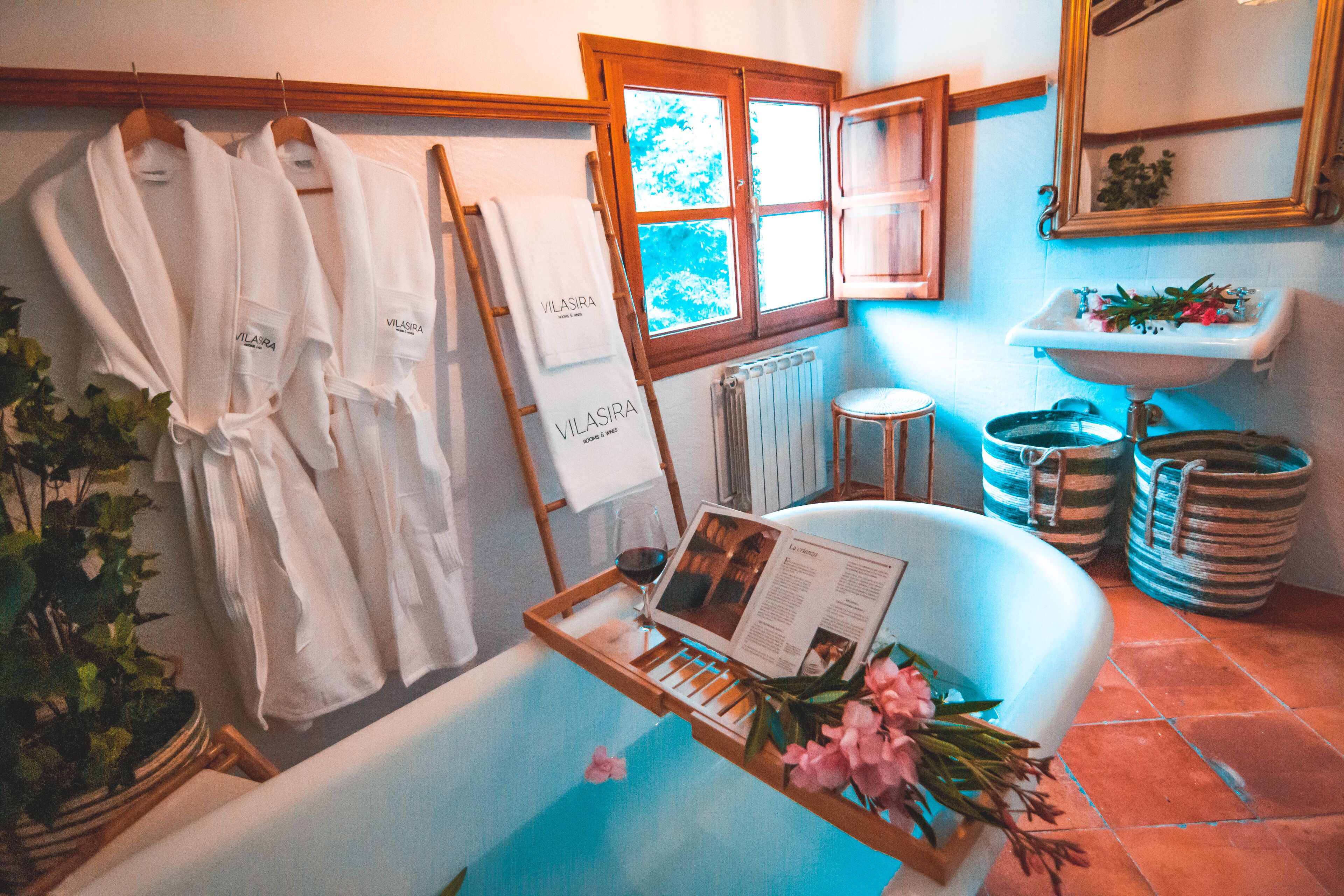 A bathroom with a bathtub, a wooden tray with a magazine and flowers, and two white robes hanging on a rack.