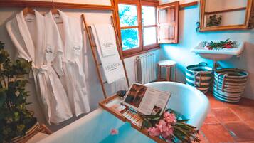 Un baño con una bañera, una bandeja de madera con una revista y flores, y dos albornoces blancos colgados en un perchero.