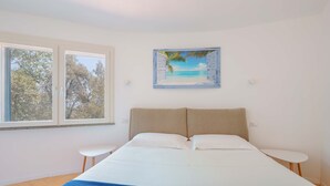 Un dormitorio con cama, ventana con vistas a árboles y un cuadro de una escena de playa.