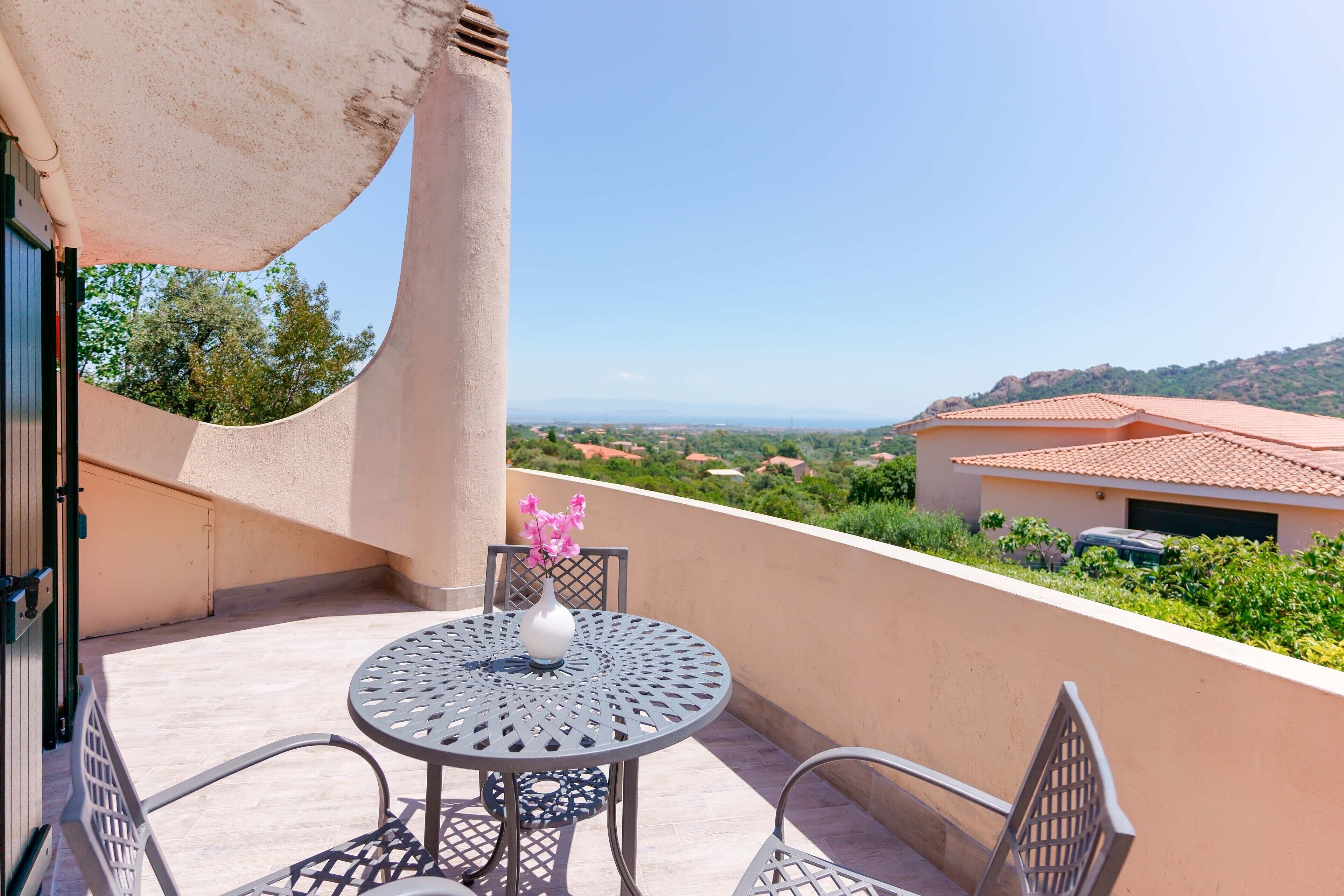 A balcony with a round table and chairs, a vase with flowers, and a view of the landscape.