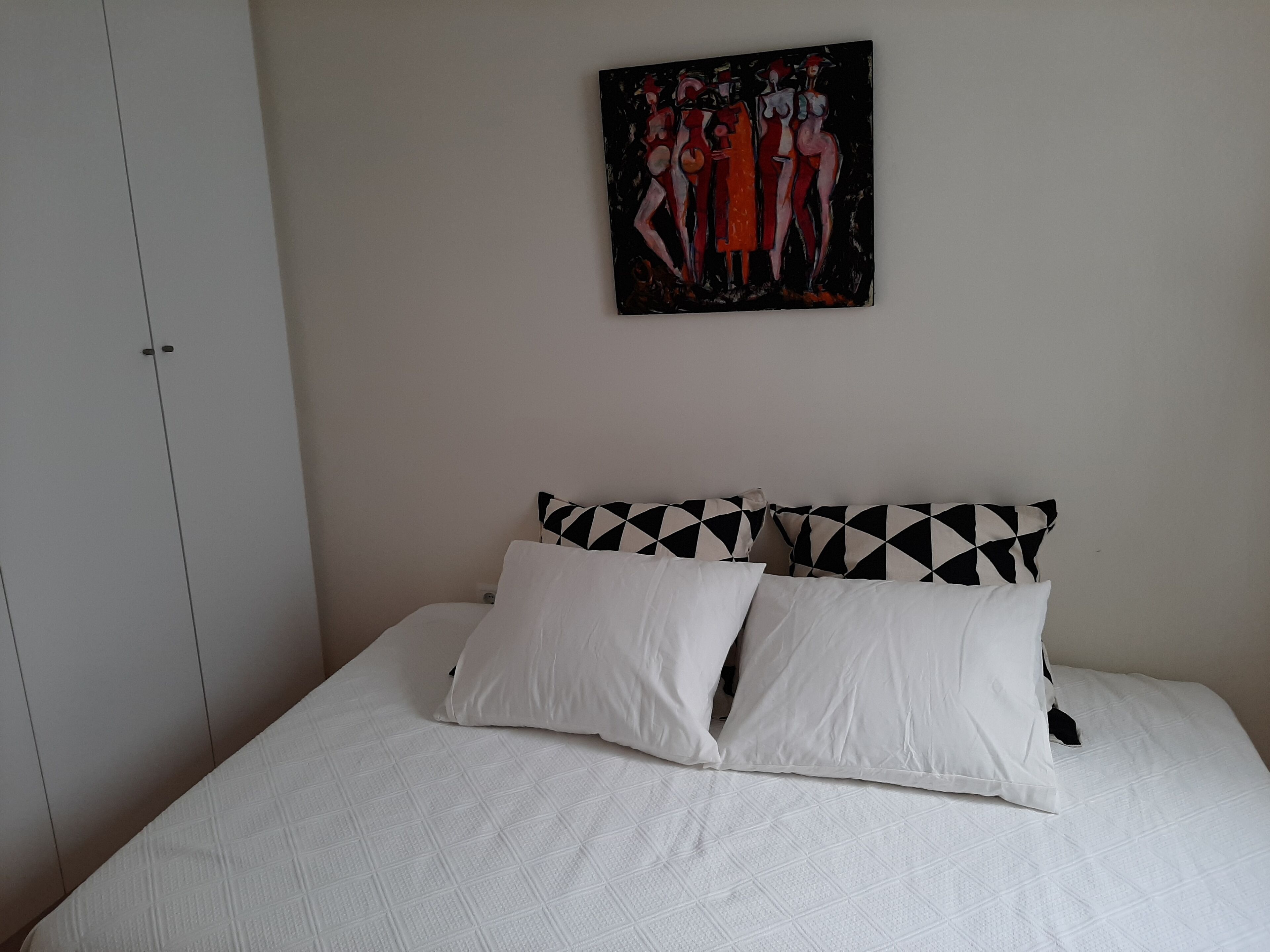 A bedroom with a bed, white bedding, a black and white patterned pillow, and a painting on the wall.