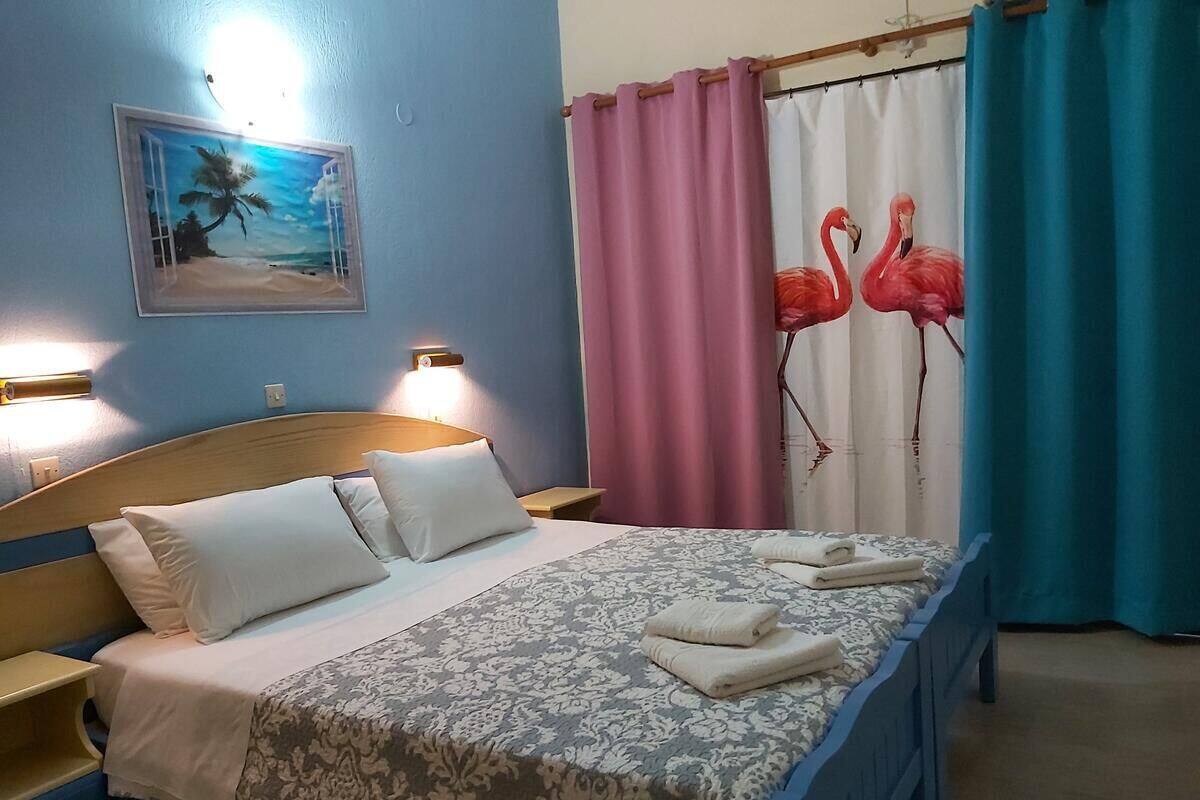 A hotel room with a bed, two bedside tables, a nightstand, a painting of a beach scene, and curtains with flamingo designs.