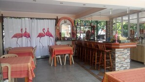 A dining area with tables and chairs, a bar with stools, and a view through large windows. - Flamingo Apartments by CorfuEscapes (Corfu)