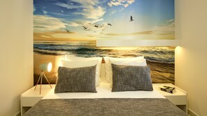Una camera da letto con un grande murale raffigurante un tramonto sull'oceano, un letto con biancheria grigia e un comodino con una lampada e un libro.