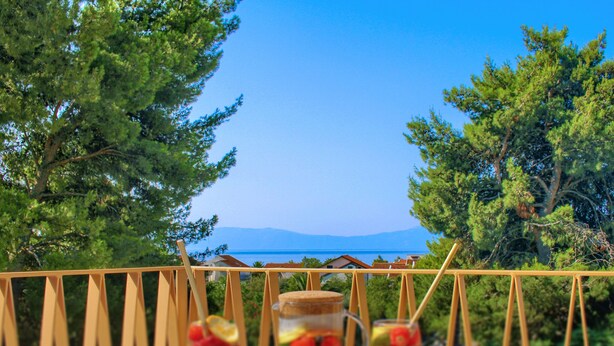 Un tavolo con due bicchieri e una caraffa, con sullo sfondo alberi e vista sul mare.