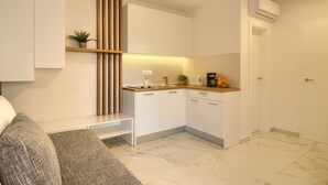 Una cucina compatta con mobili bianchi, uno scaffale in legno con una pianta, un pavimento in marmo e un piccolo bancone con una macchina da caffè e arance.