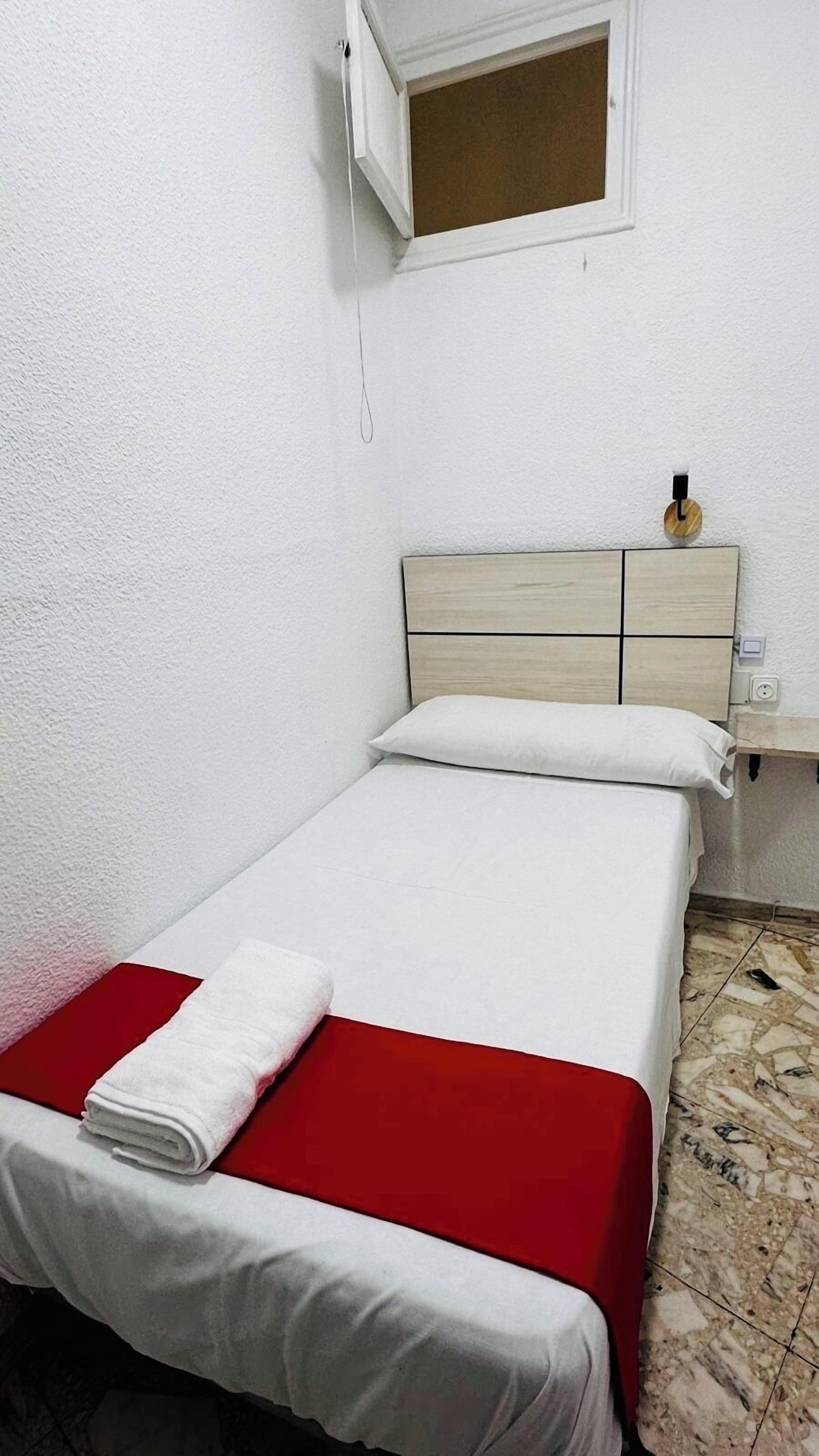 A small, neatly made bed with a red and white bedspread, a white pillow, and a rolled white towel on the footboard.