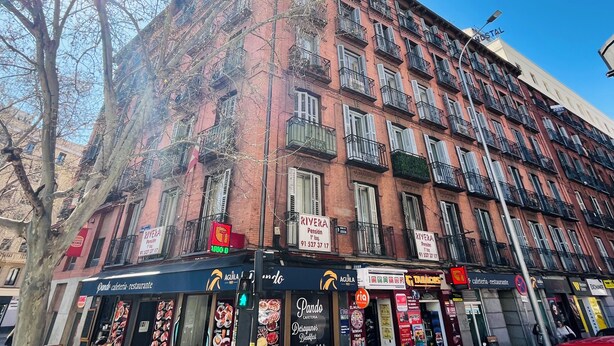 A multi-story building with a red brick facade, multiple balconies, and various commercial establishments on the ground floor. - Pension Rivera (Madrid)