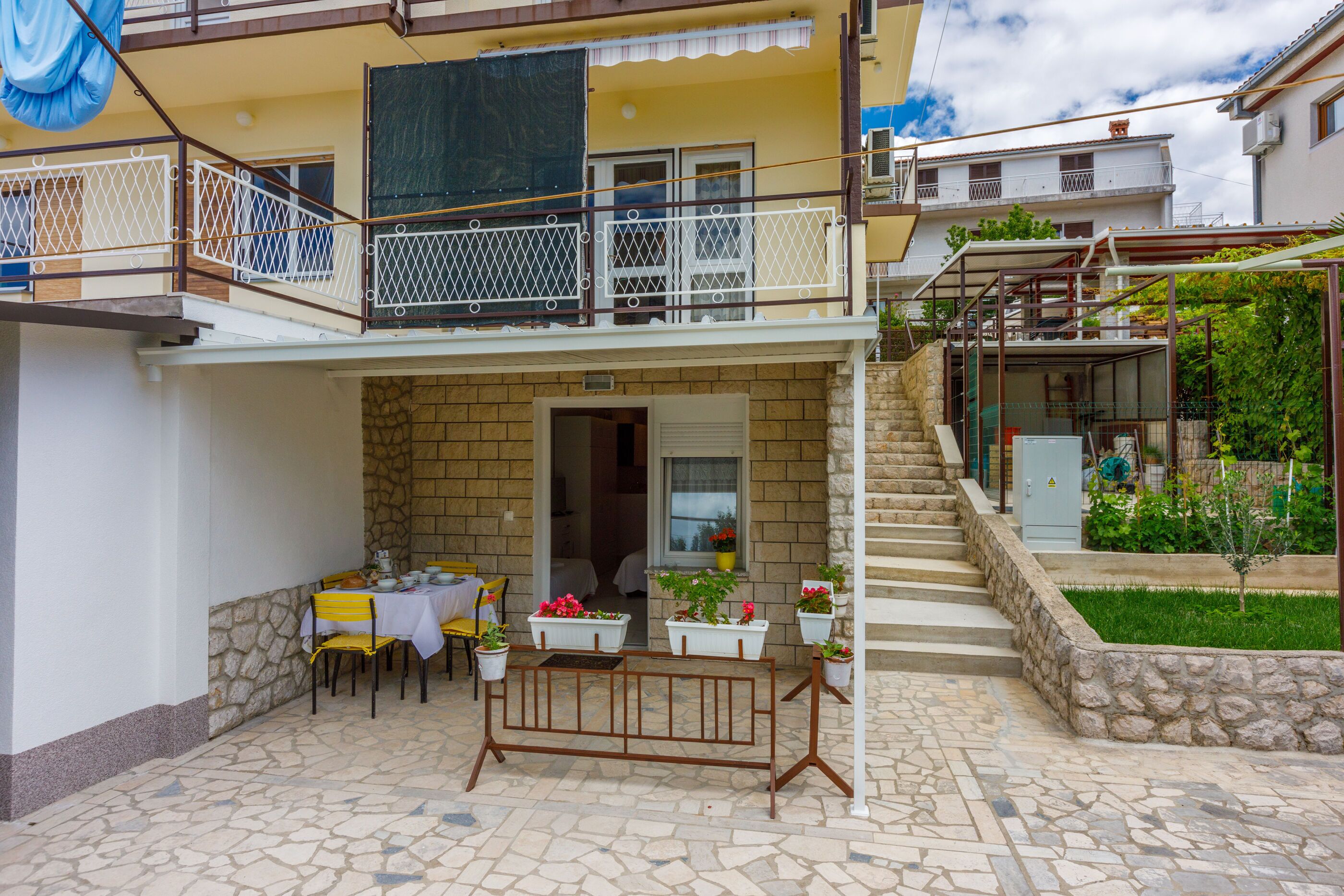 Ein zweistöckiges Gebäude mit Balkon, Naturstein-Fassade, kleiner Außenbereich mit Tisch und Stühlen sowie Topfpflanzen.