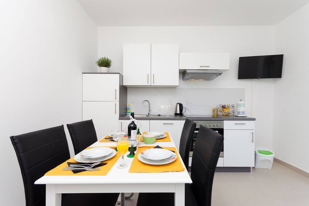 A modern kitchen with white cabinets, a flat-screen TV mounted above the counter, and a dining area with a table set for two.