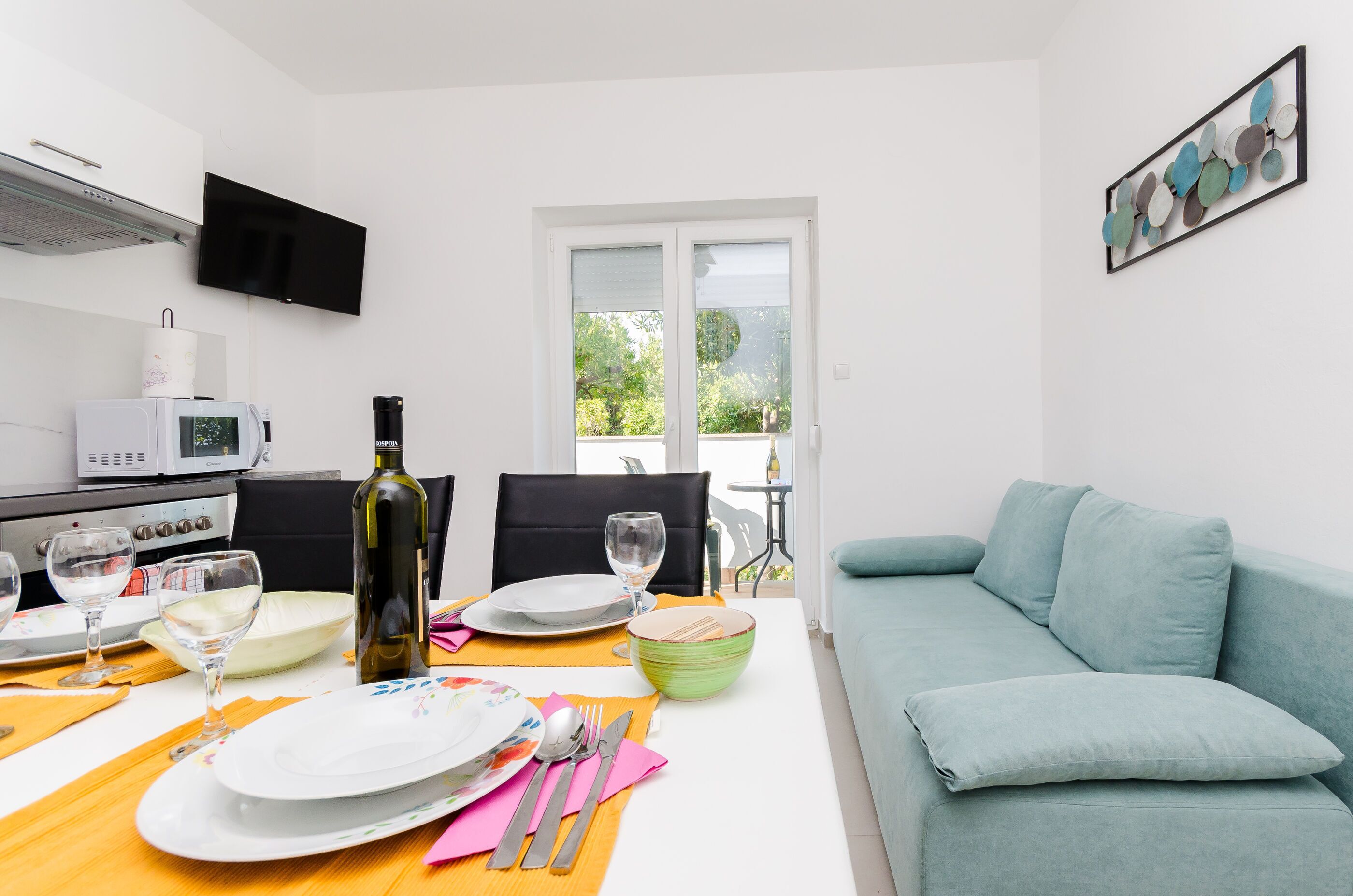 A modern kitchen with a dining area, a TV mounted on the wall, and a view of the outdoors through a large window.