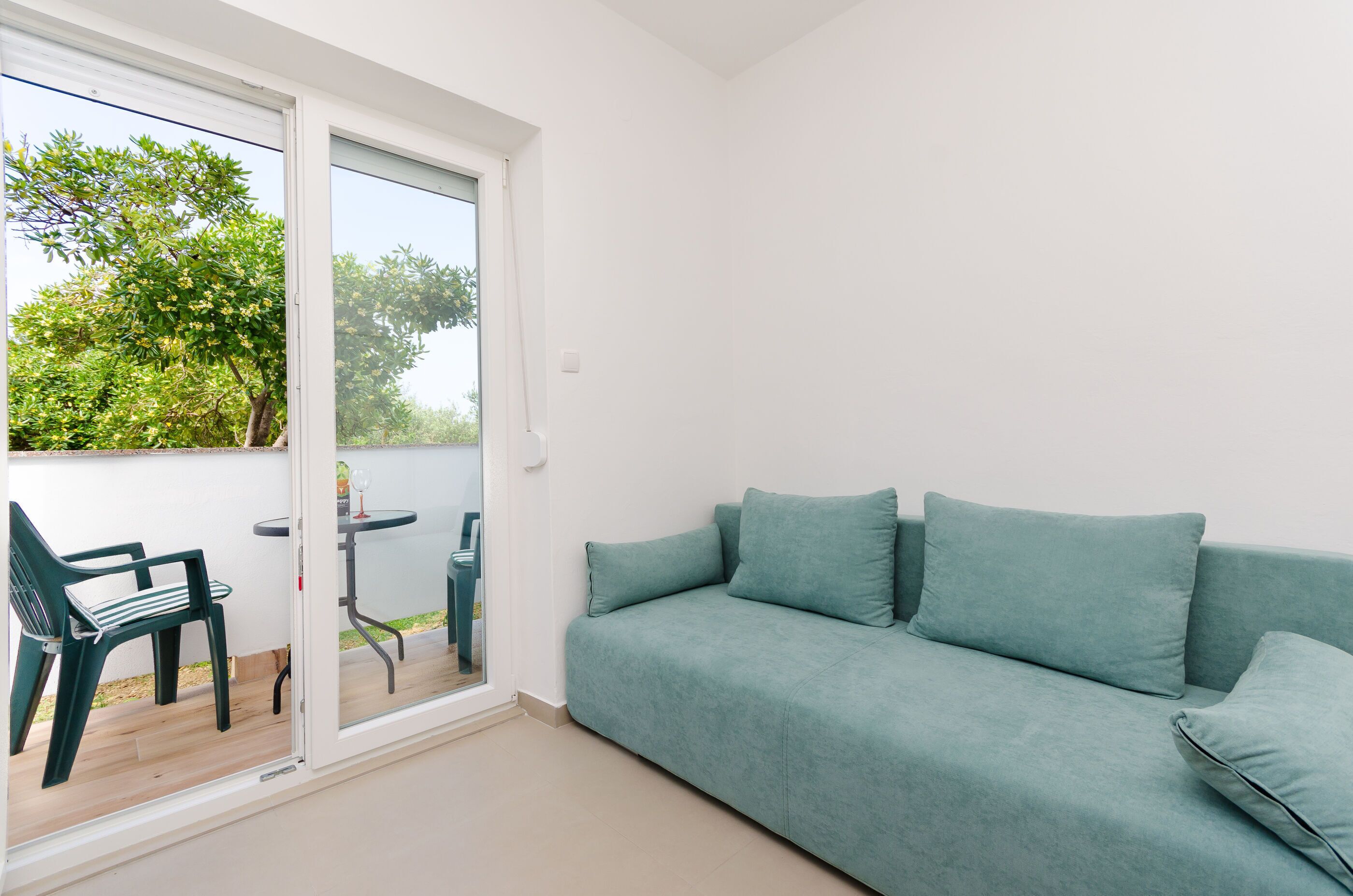A room with a teal sofa, a small table, and a chair near a sliding glass door leading to an outdoor area with greenery.