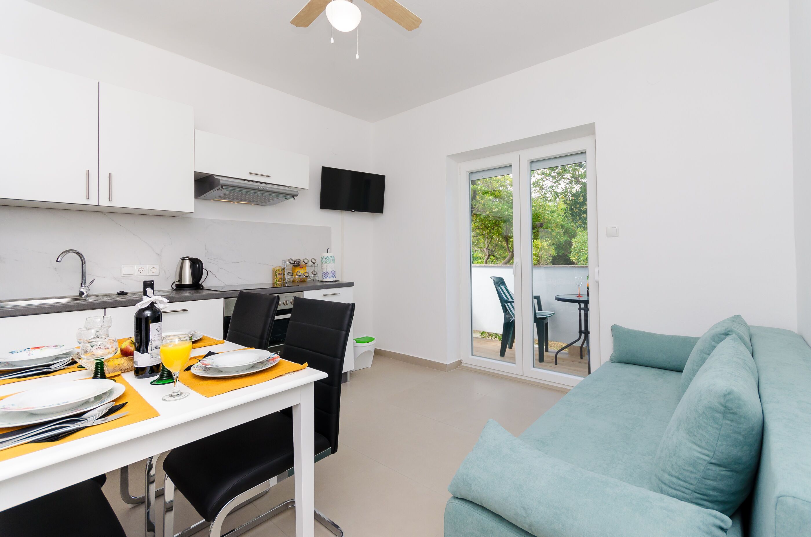 A modern kitchen with white cabinets, a dining table set for two, and a sofa in the living area.