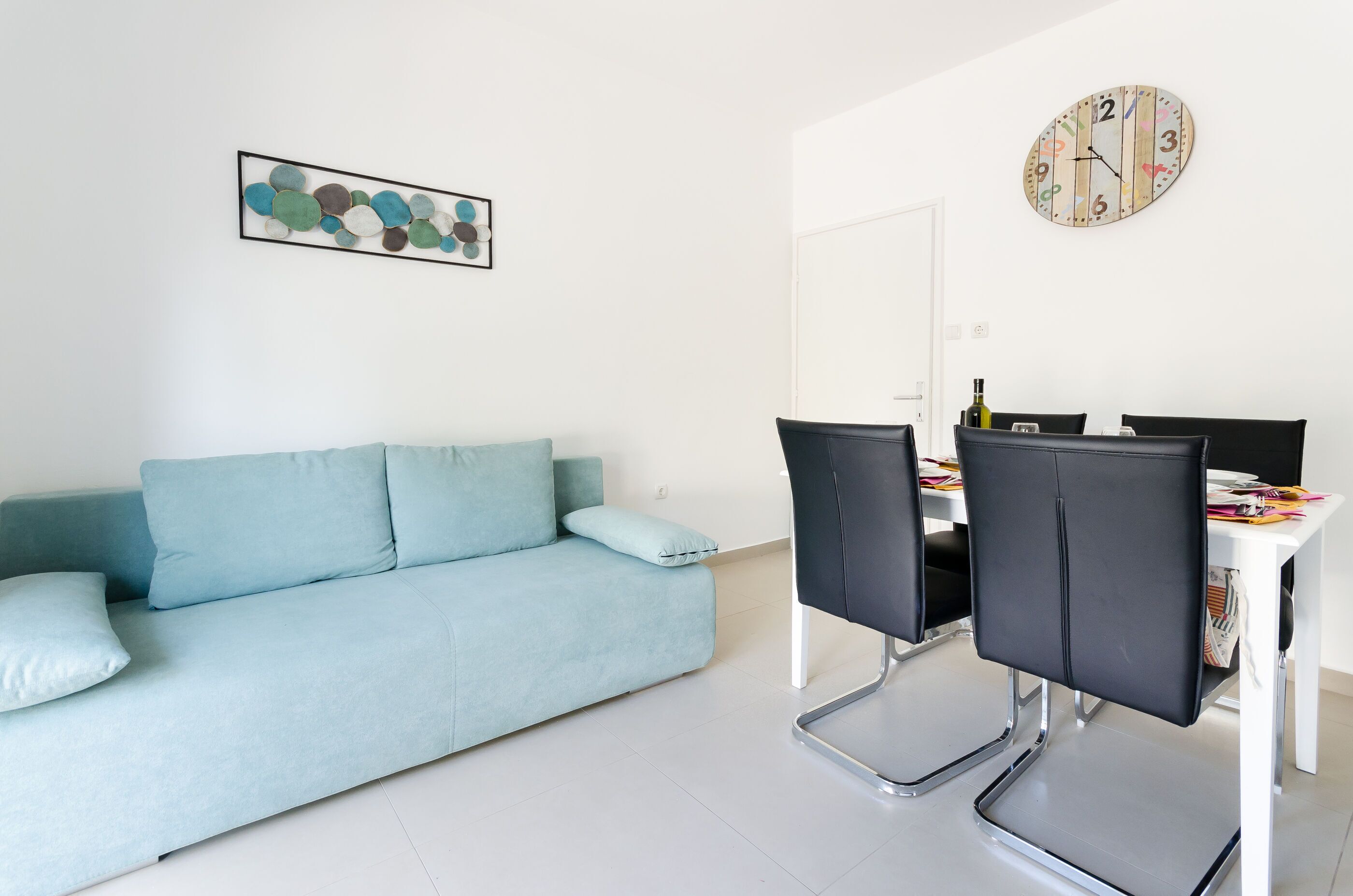 A modern living room with a light blue sofa, a dining table with chairs, a clock, and a framed abstract art piece.