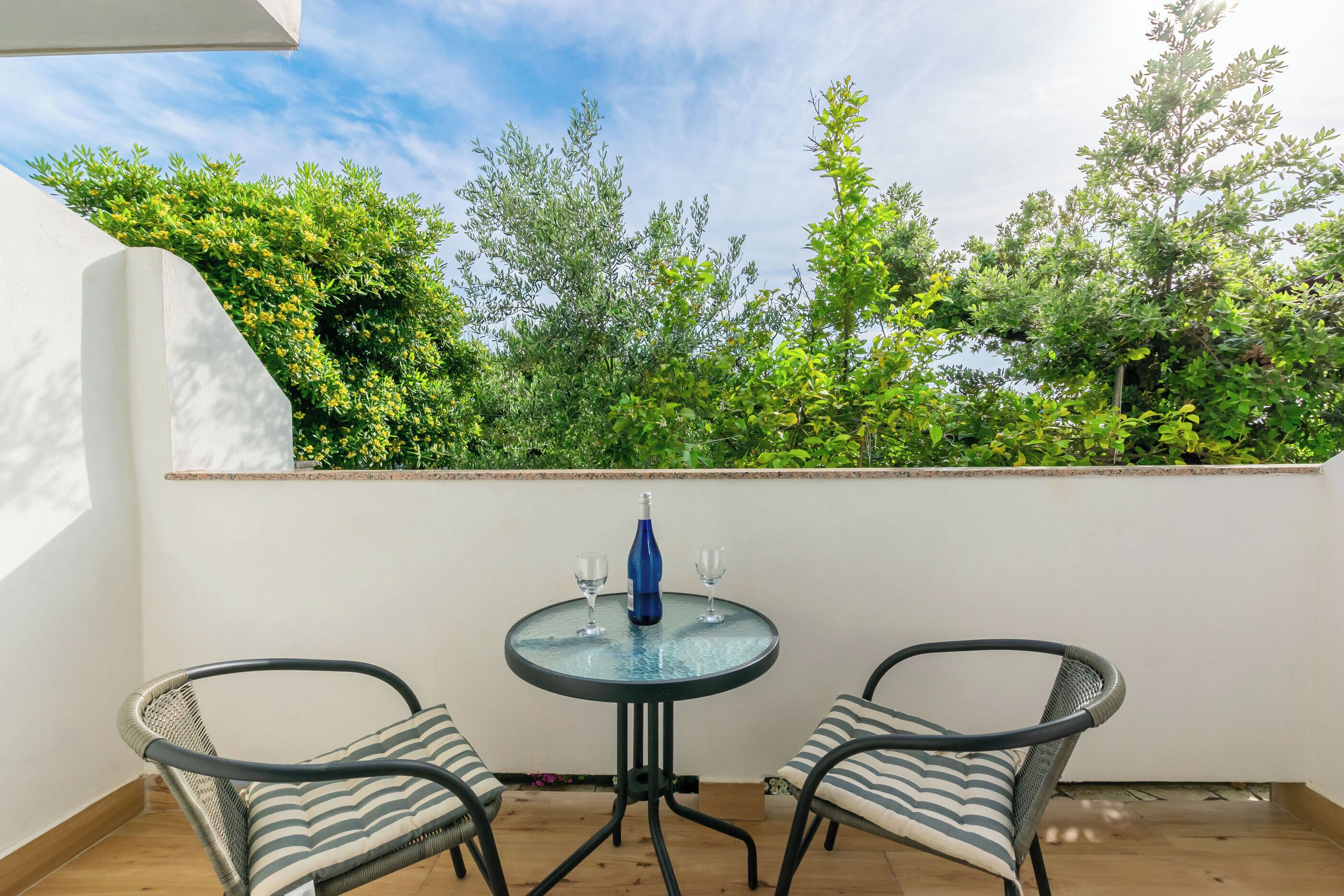 Un piccolo patio esterno con un tavolo rotondo in vetro, due sedie in vimini e una bottiglia d'acqua.