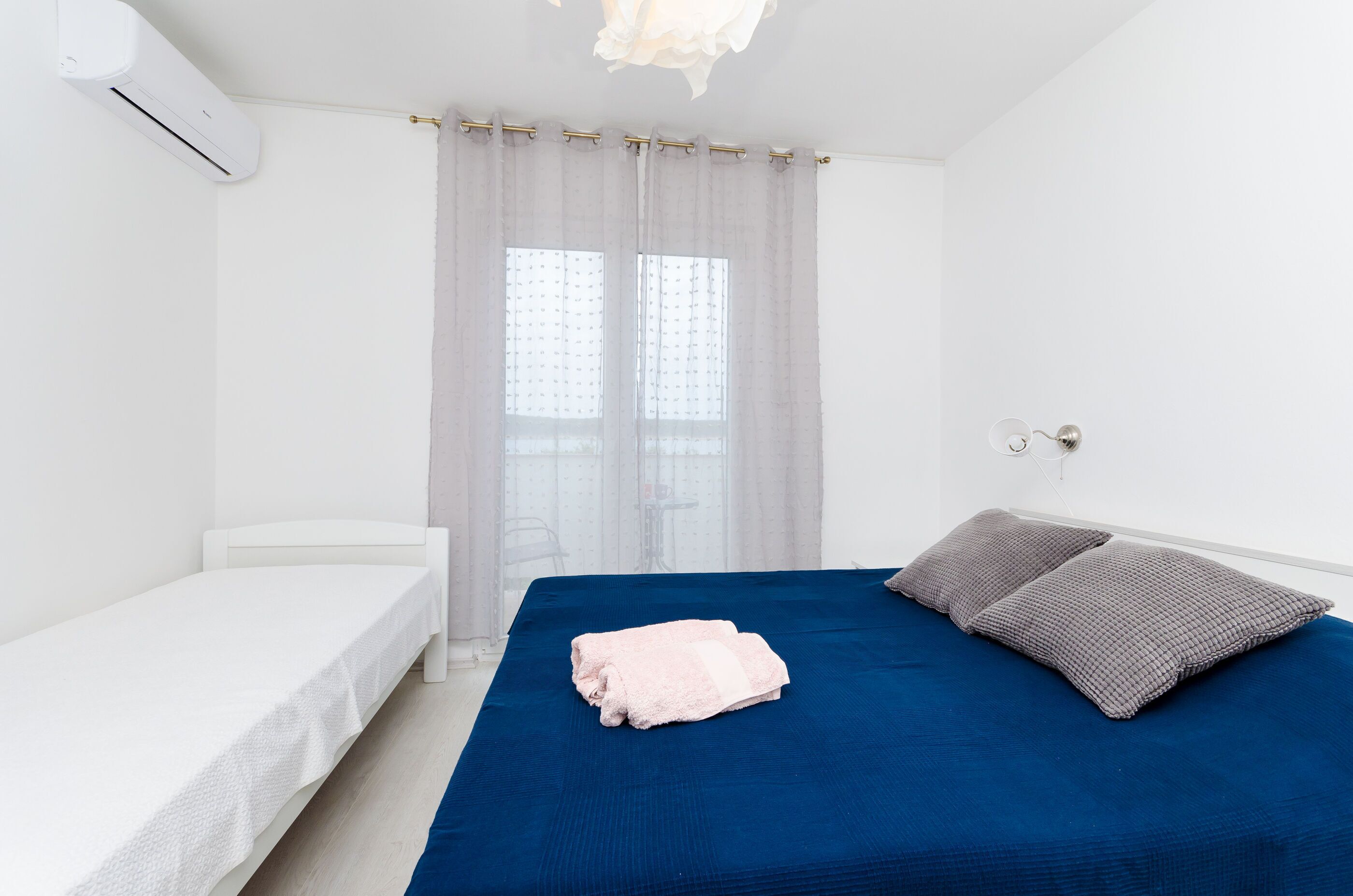 A bedroom with a single bed, a smaller bed, a window with sheer curtains, and a wall-mounted air conditioning unit.