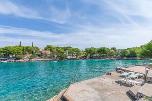 A clear turquoise pool with a concrete deck, lounge chairs, and a stone staircase leading into the water. - Apartment Vesna (Milna)