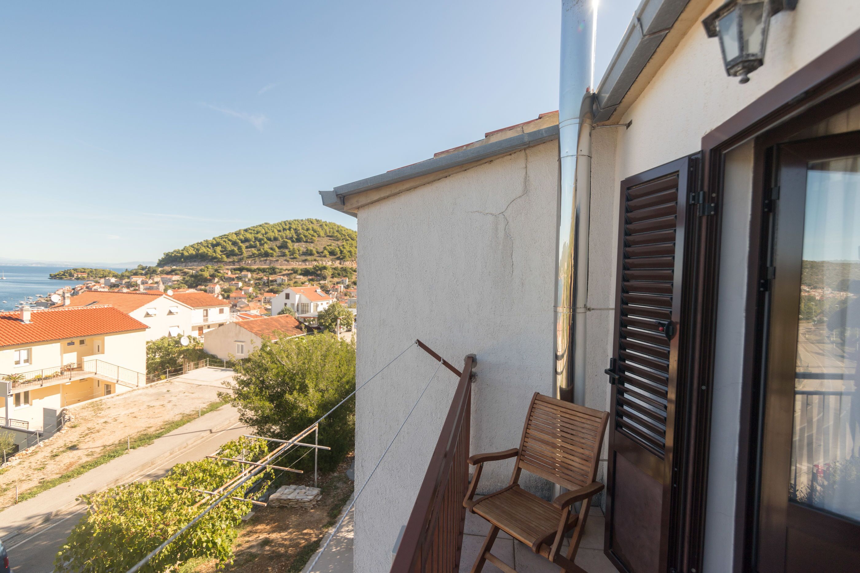 A balcony with a wooden chair, overlooking a coastal town with red-roofed buildings and a hillside.