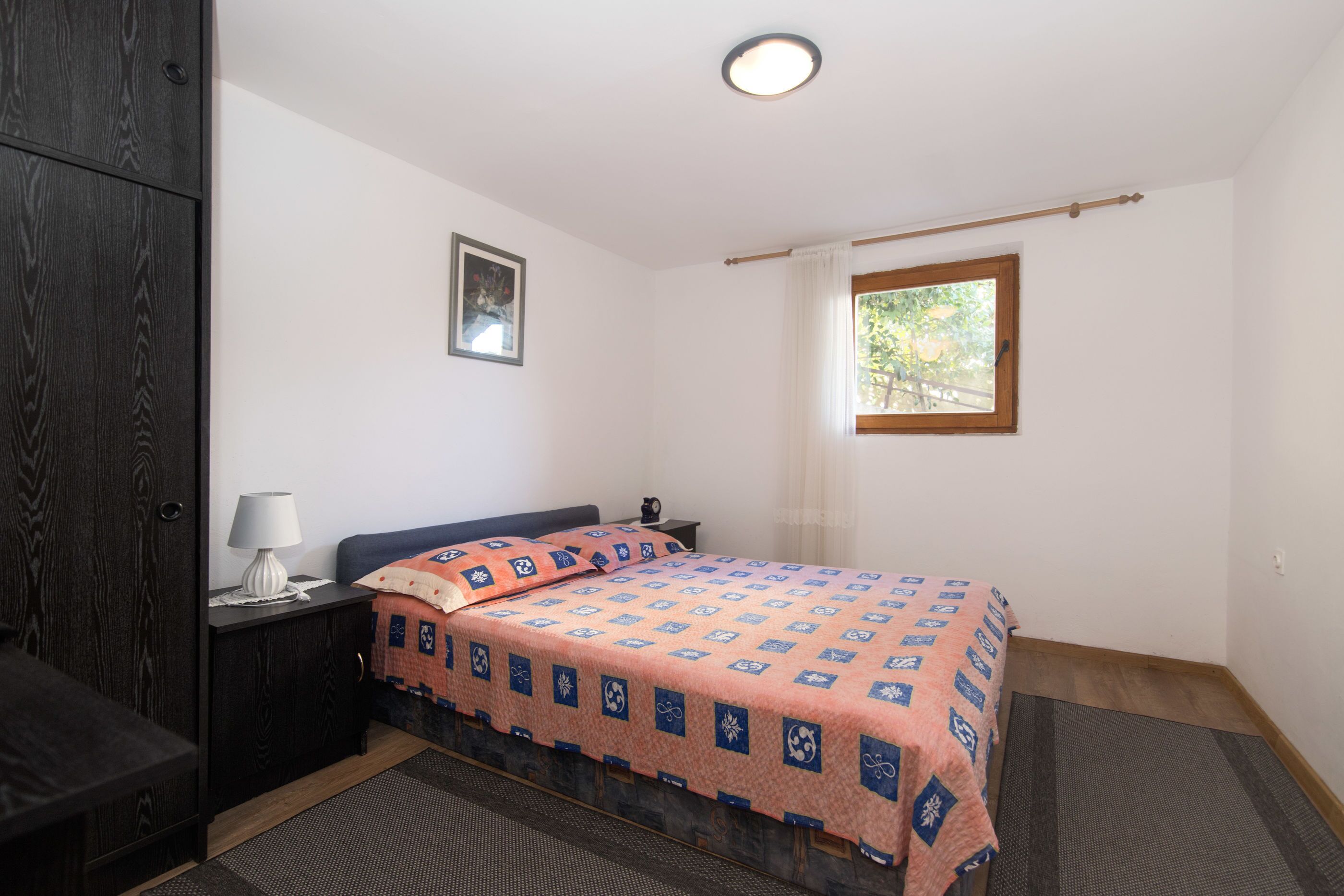 A bedroom with a bed, a nightstand, a lamp, a dark wardrobe, a window with a view, and a framed picture on the wall.