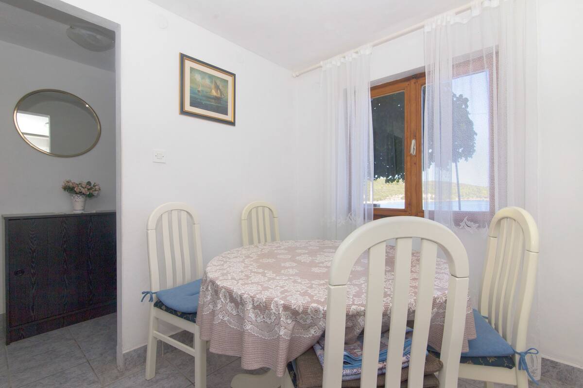 Un comedor con una mesa redonda cubierta por un mantel de encaje, cuatro sillas blancas con cojines azules, un cuadro enmarcado en la pared y una ventana con cortinas traslúcidas.