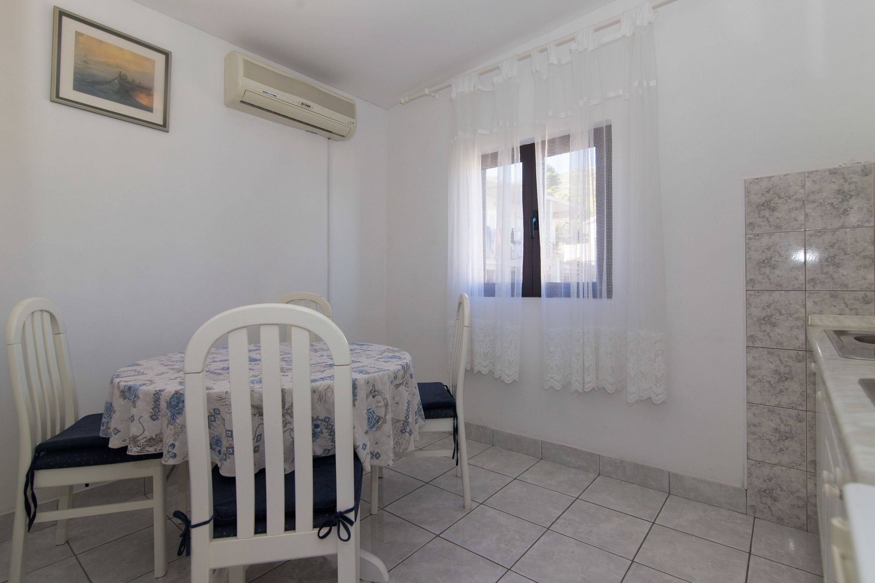 A dining area with a table and chairs, a window with sheer curtains, and an air conditioning unit on the wall.
