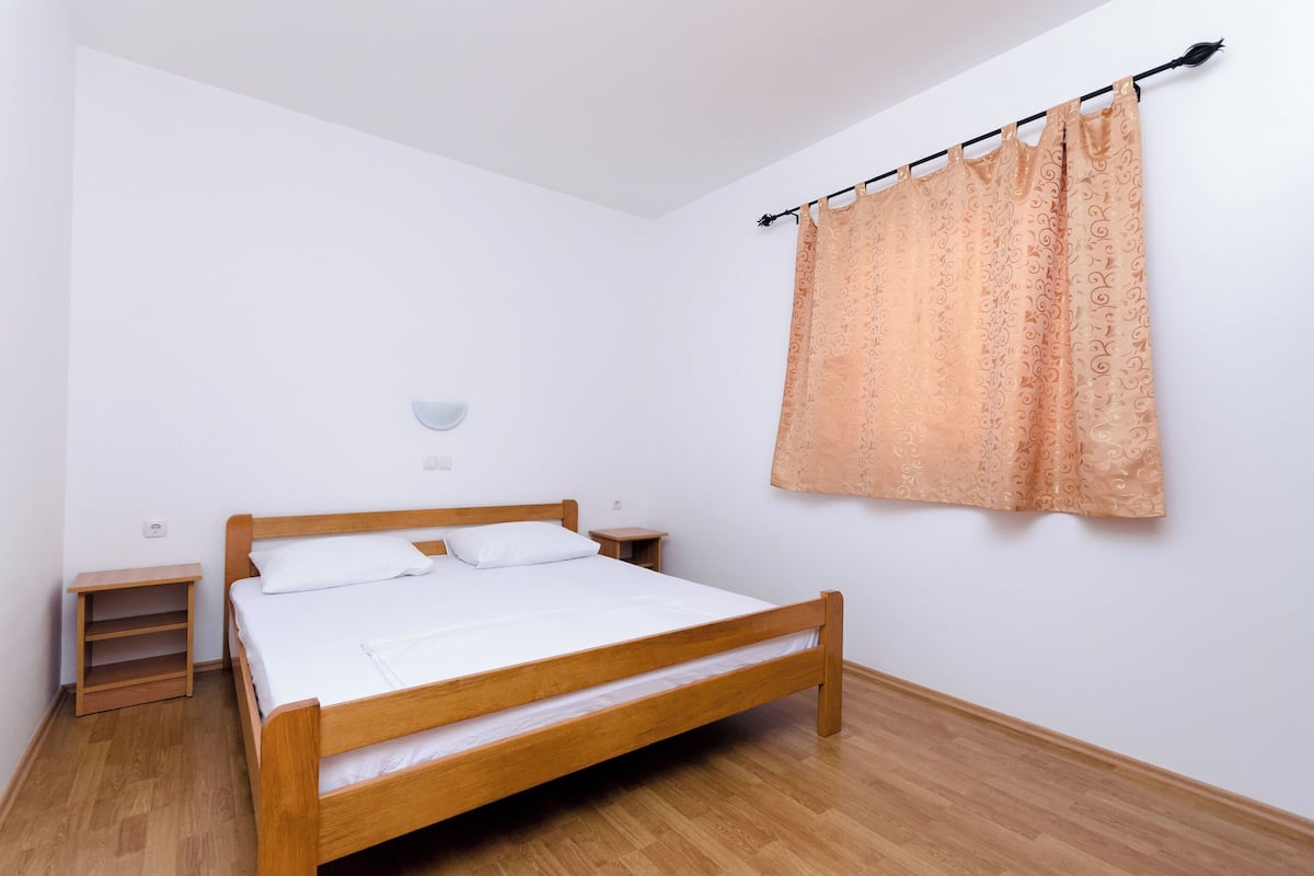 A bedroom with a wooden bed, white bedding, a nightstand, a small shelf, and a patterned curtain.