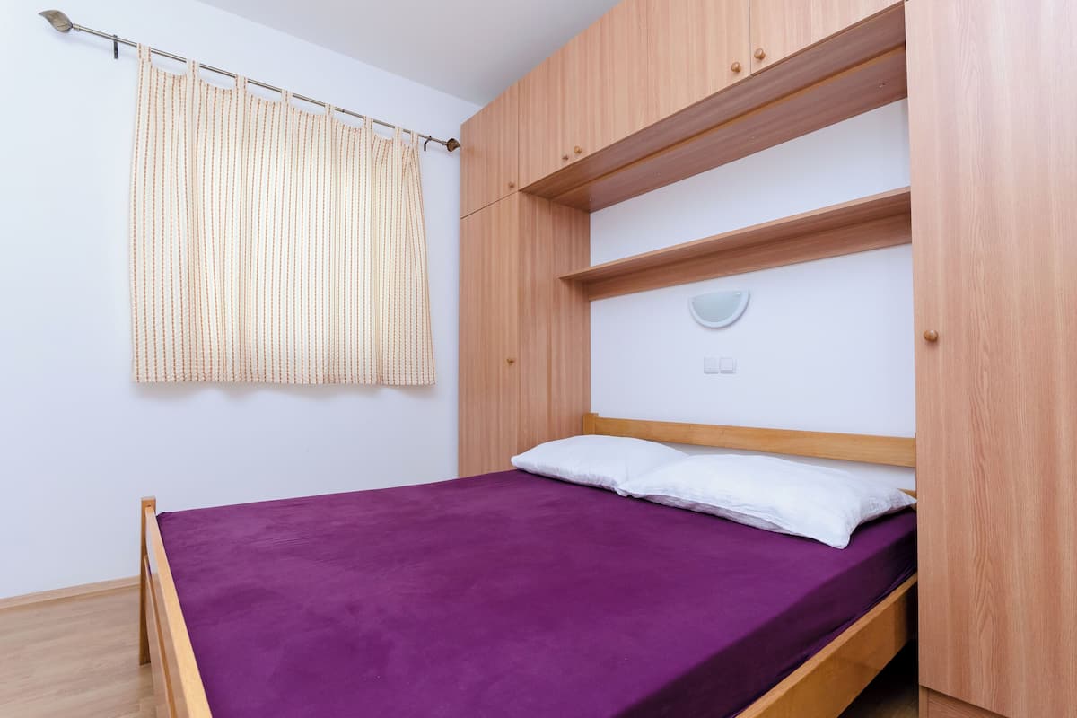 A single bed with a purple mattress and white pillows, a wooden headboard, and a window with a curtain.