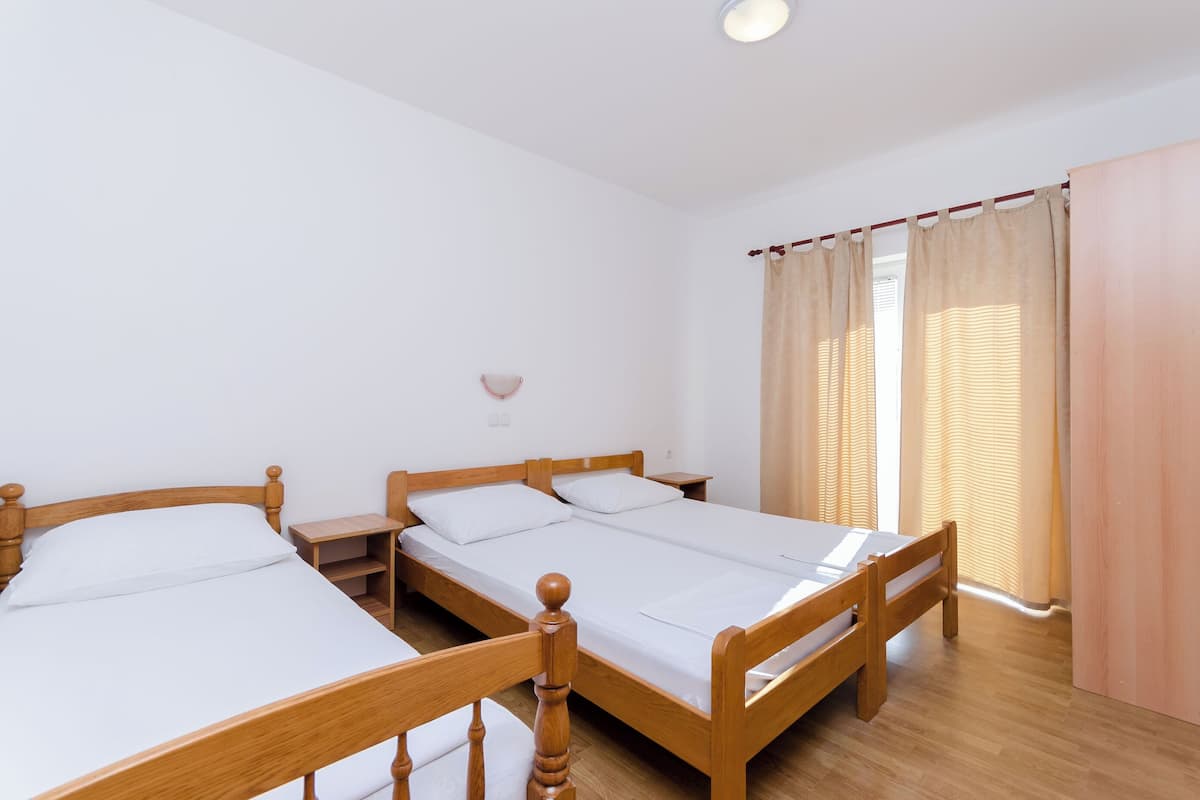 A room with two wooden beds, white bedding, a small bedside table, a wall-mounted light, and a window with curtains.