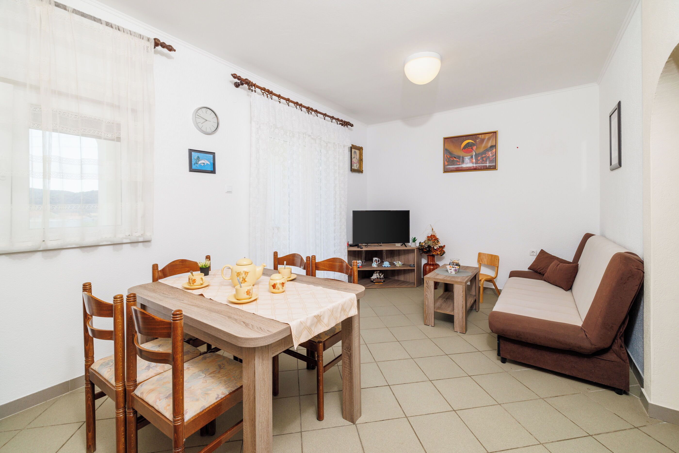 A living room with a wooden dining table, chairs, a sofa, a TV, and a small table with decorative items.