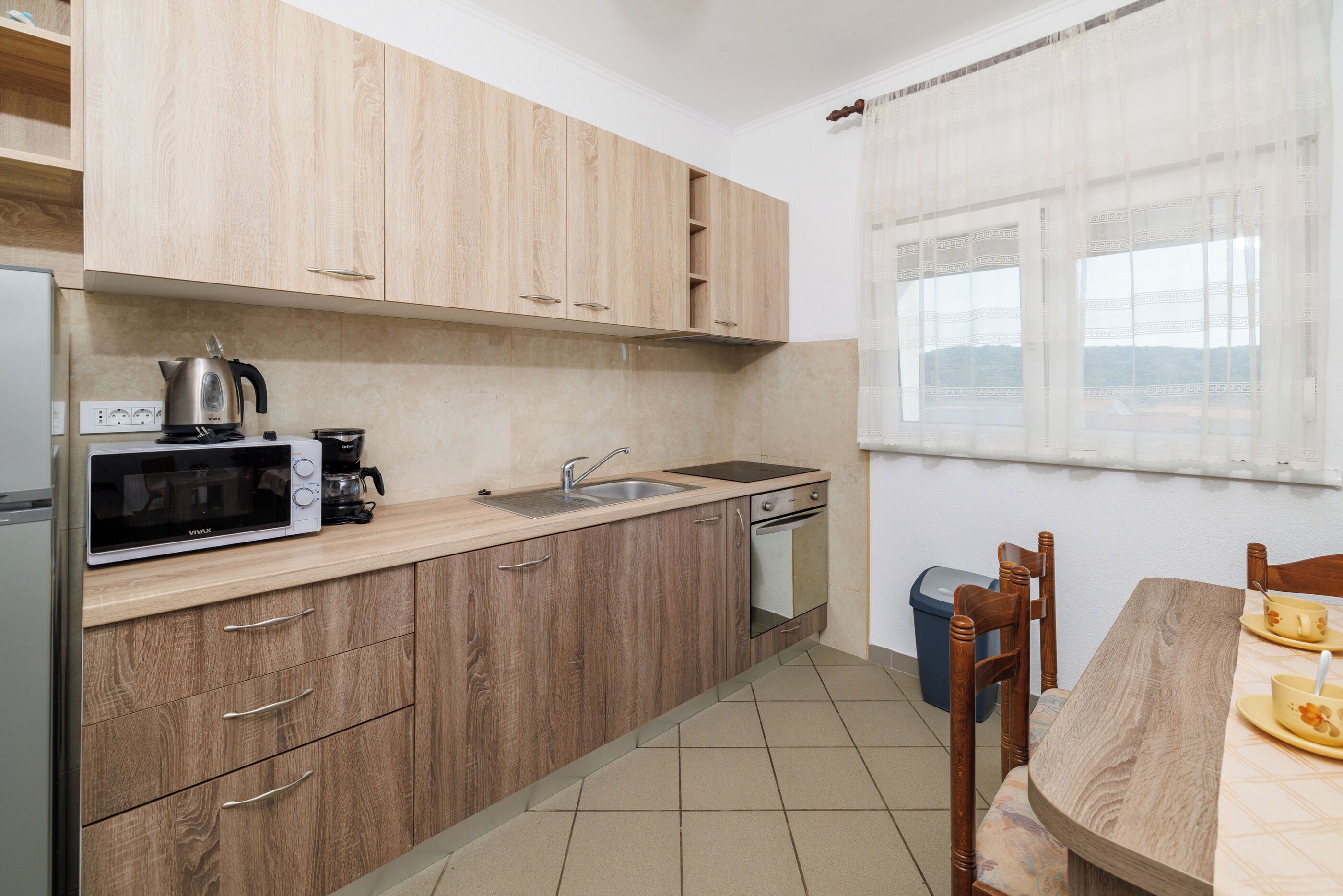A kitchen with wooden cabinets, a microwave, a kettle, a sink, and a dining table with chairs.