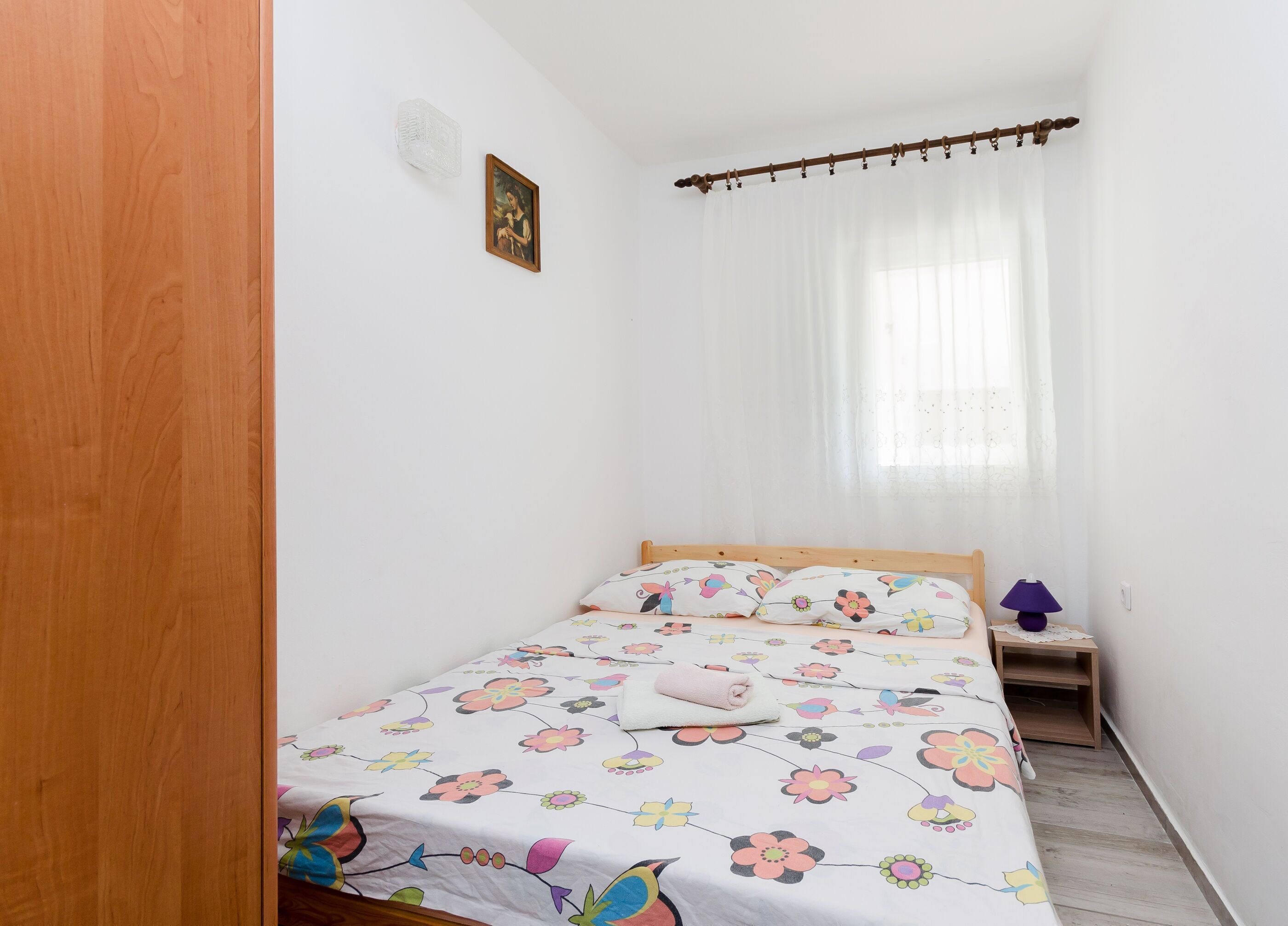 A small, neatly arranged bedroom with a single bed, a bedside table, a lamp, and a window with sheer curtains.