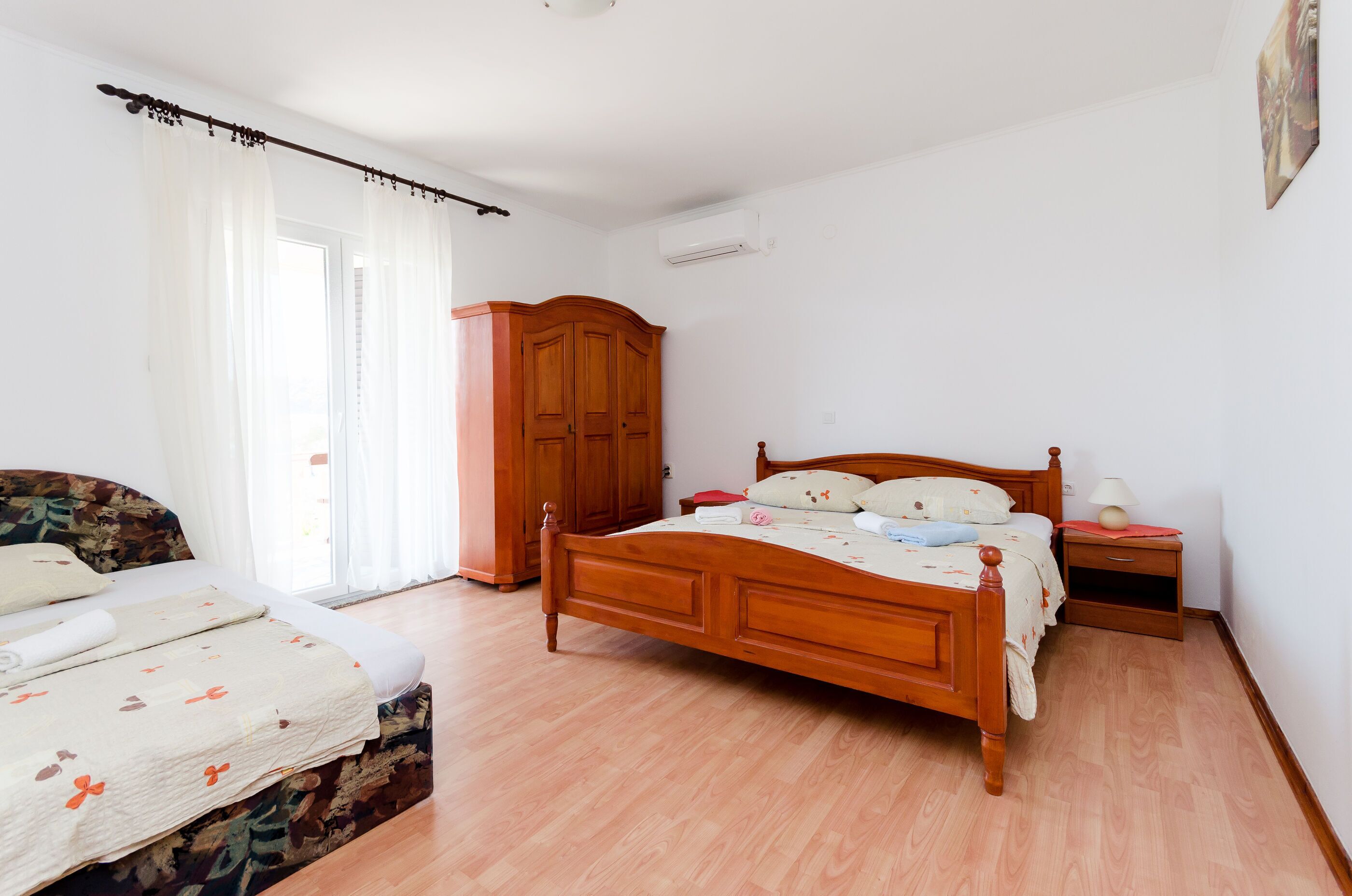 A bedroom with a wooden bed, a wooden wardrobe, a bedside table with a lamp, and a window with sheer curtains.