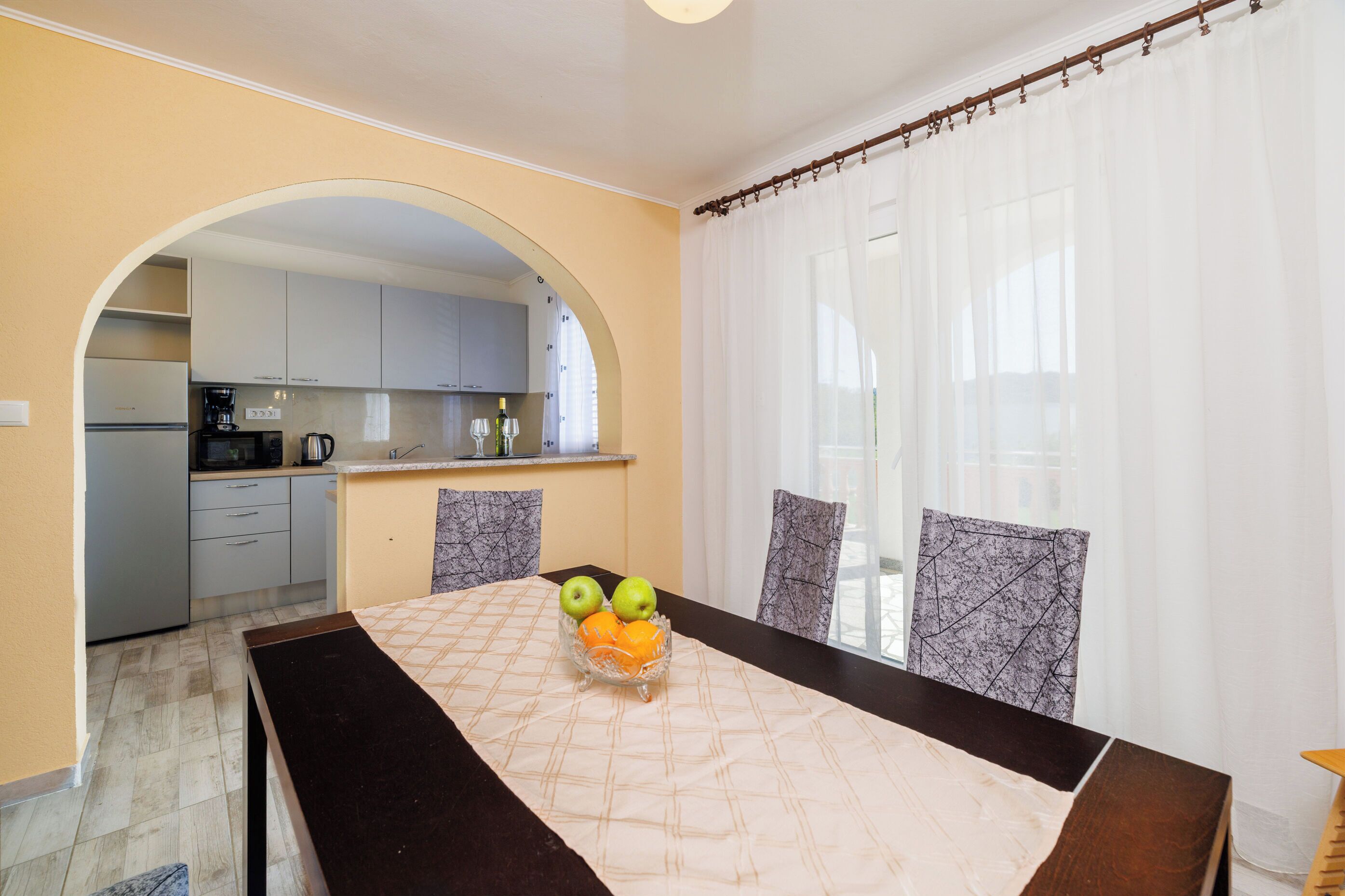 Een eethoek met een tafel gedekt voor twee personen, een keuken op de achtergrond en een uitzicht door een raam met dunne gordijnen.