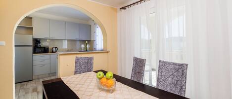 A dining area with a table set for two, a kitchen in the background, and a view through a window with sheer curtains.