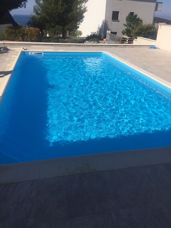 A rectangular swimming pool with clear blue water, surrounded by a tiled deck and a building in the background. - Apartments Lanterna (Rogoznica)