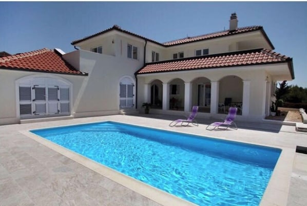 Une maison à deux étages avec une piscine, un patio couvert doté de chaises longues et un toit en tuiles.