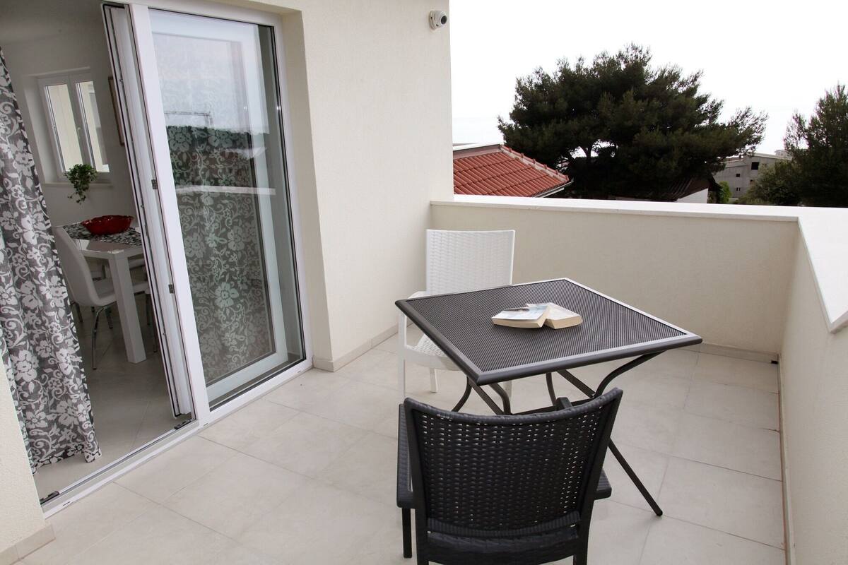 Un balcon avec une table et des chaises, une porte-fenêtre coulissante et une vue sur les arbres et les bâtiments.