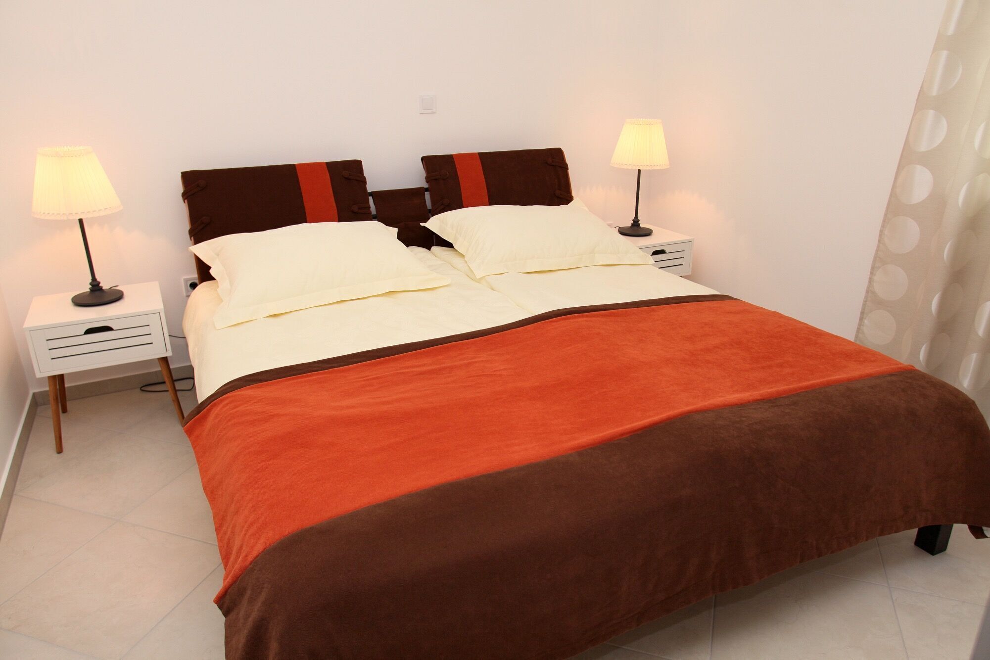 A neatly made bed with a brown and red blanket, two bedside lamps, and a white nightstand.