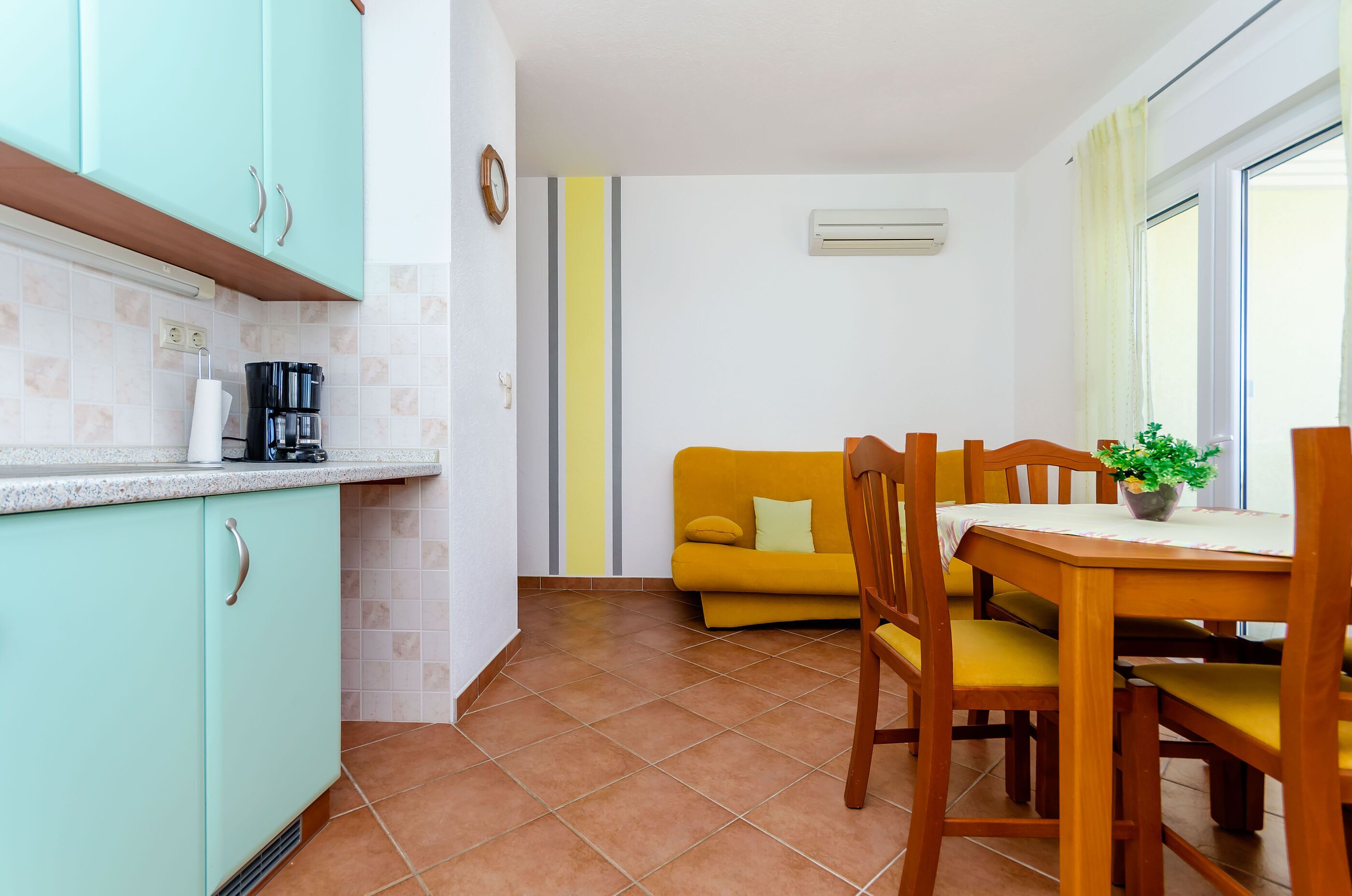A compact kitchen with teal cabinets, a coffee maker, and a yellow sofa in the adjacent living area.