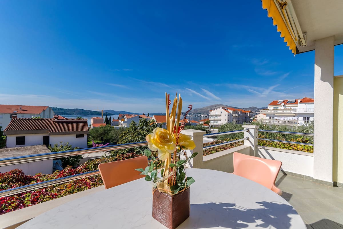 A balcony with a table and chairs, a vase with flowers, and a view of buildings and hills.