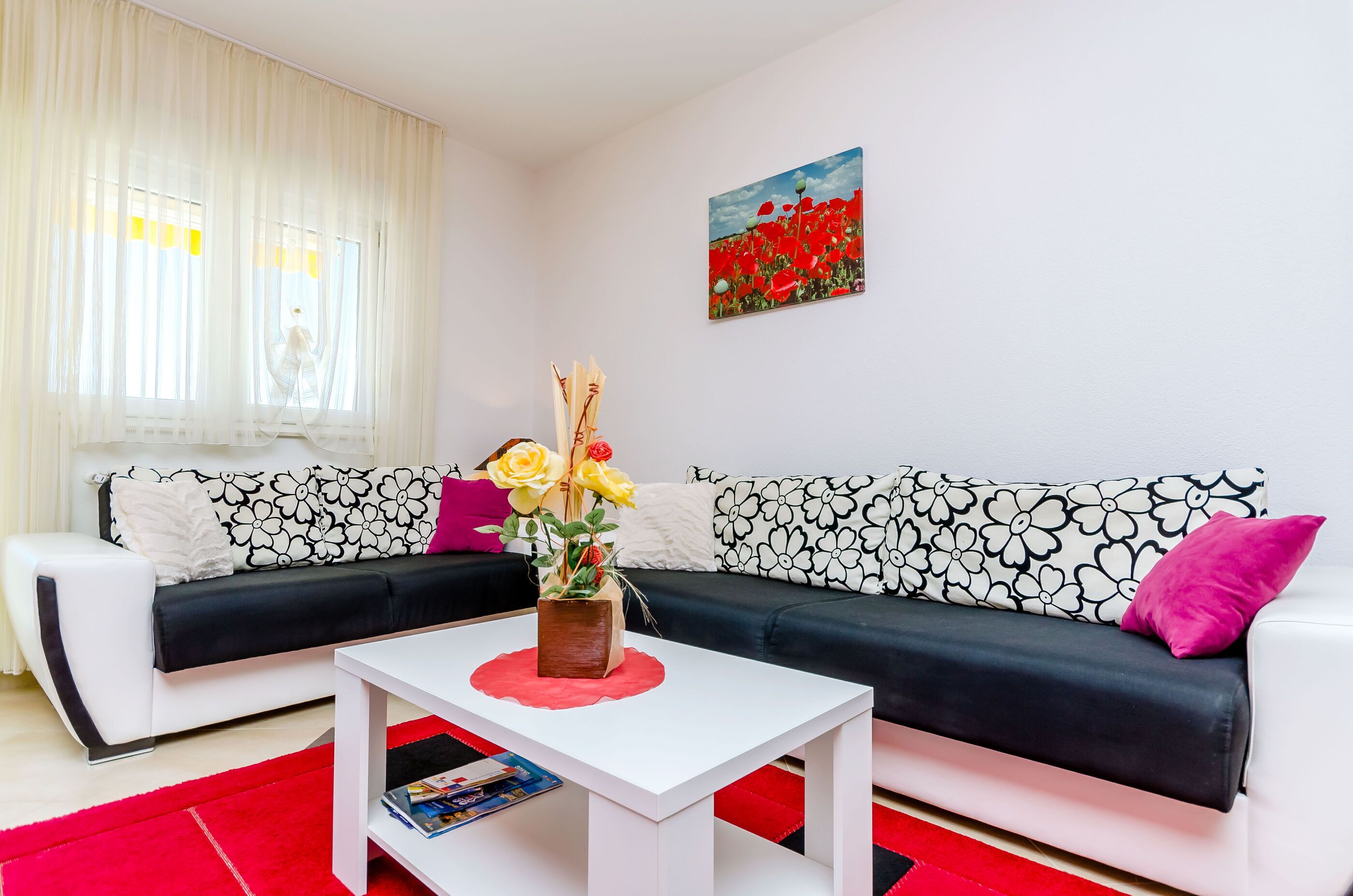 A living room with a white sofa, a black sofa, a white coffee table, and a red rug. There is a painting of red flowers on the wall.