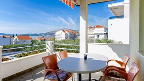 Un balcon avec une table et des chaises, donnant sur une ville côtière, avec des bâtiments et des collines en arrière-plan.