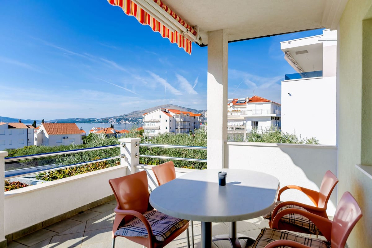 A balcony with a table and chairs, overlooking a coastal town with buildings and hills.
