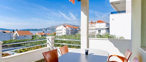 A balcony with a table and chairs, overlooking a coastal town with buildings and hills in the background.