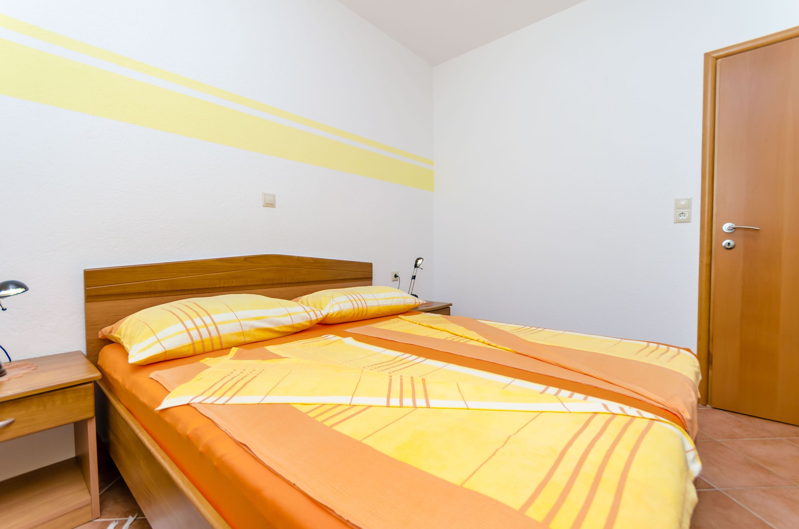 A bedroom with a wooden bed, yellow and orange bedding, a nightstand, and a wooden door.