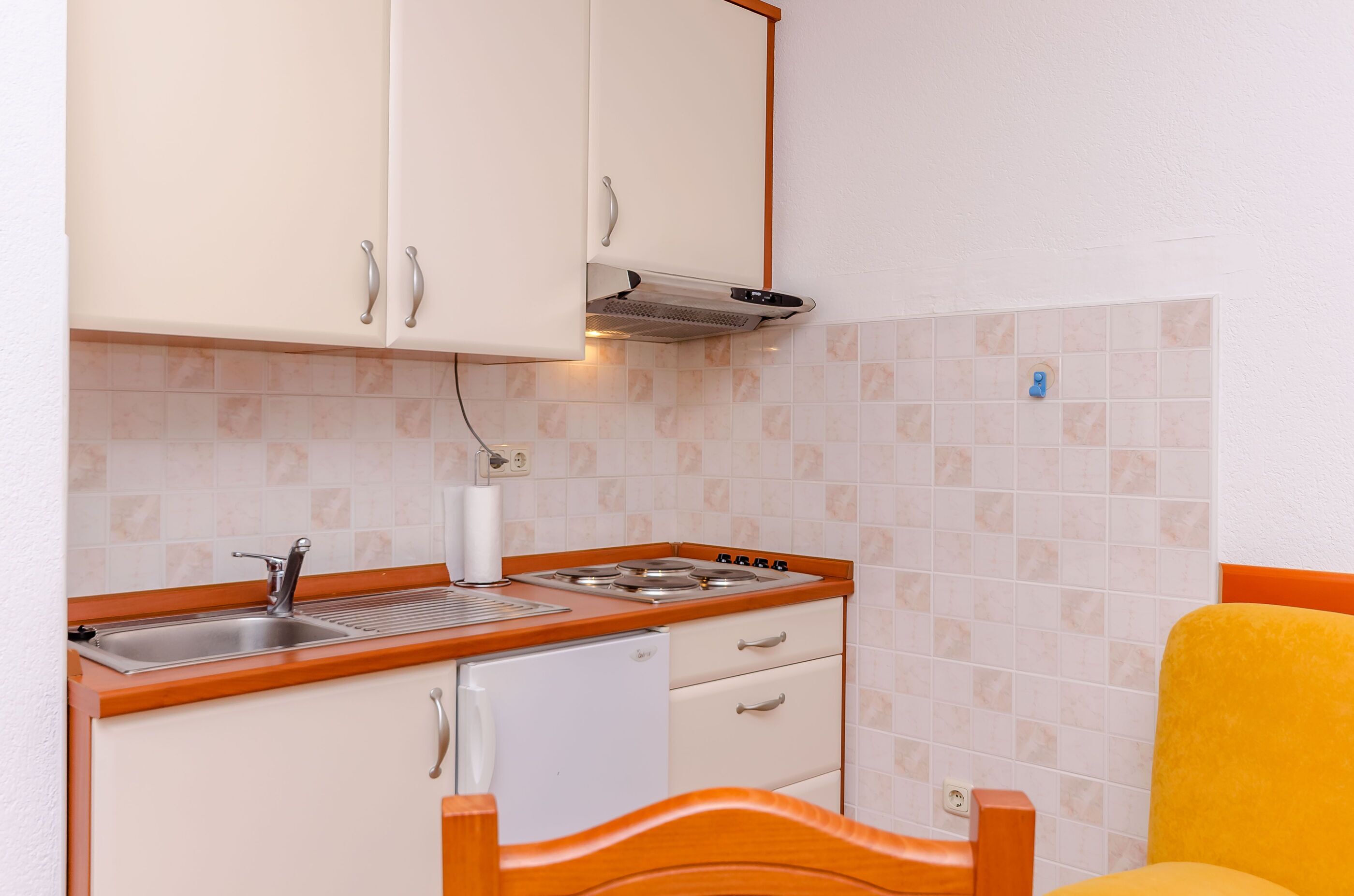 Cozinha compacta com pia, fogão e geladeira branca. Há uma cadeira de madeira com almofada amarela.