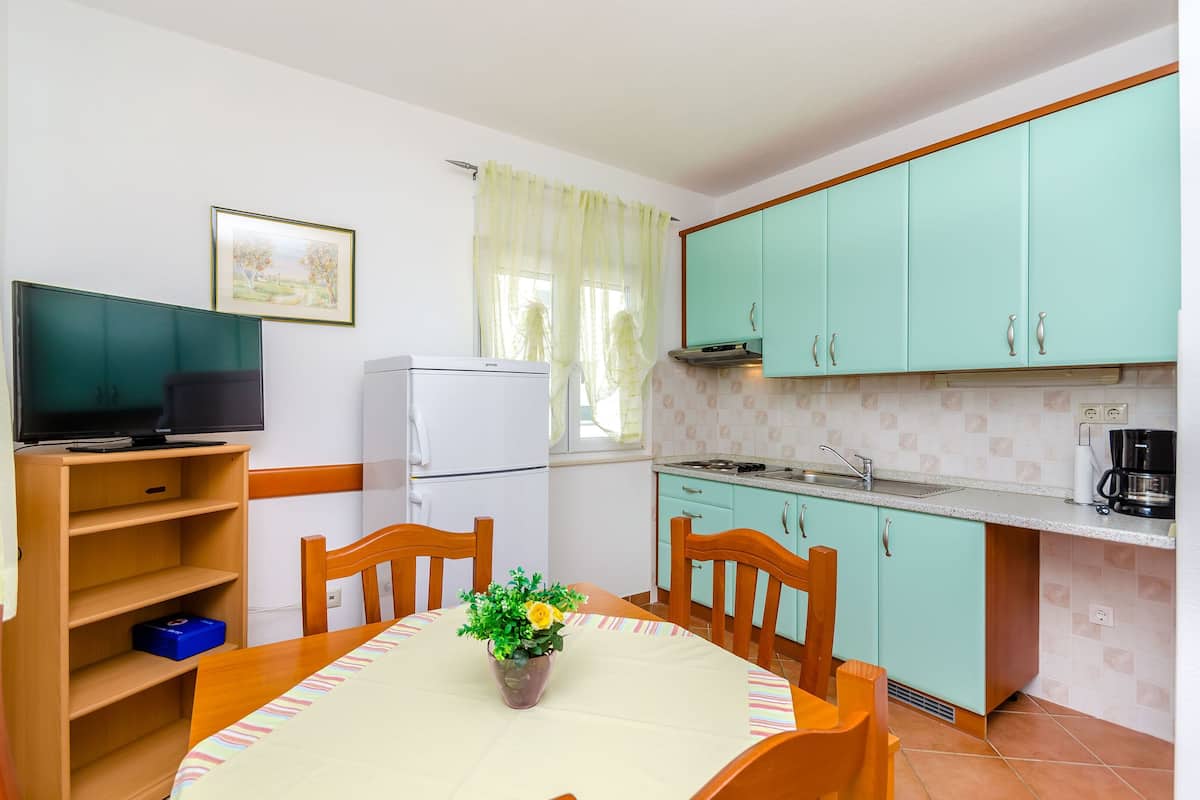 A compact kitchen with teal cabinets, a white refrigerator, a dining table with chairs, and a TV on a wooden stand.