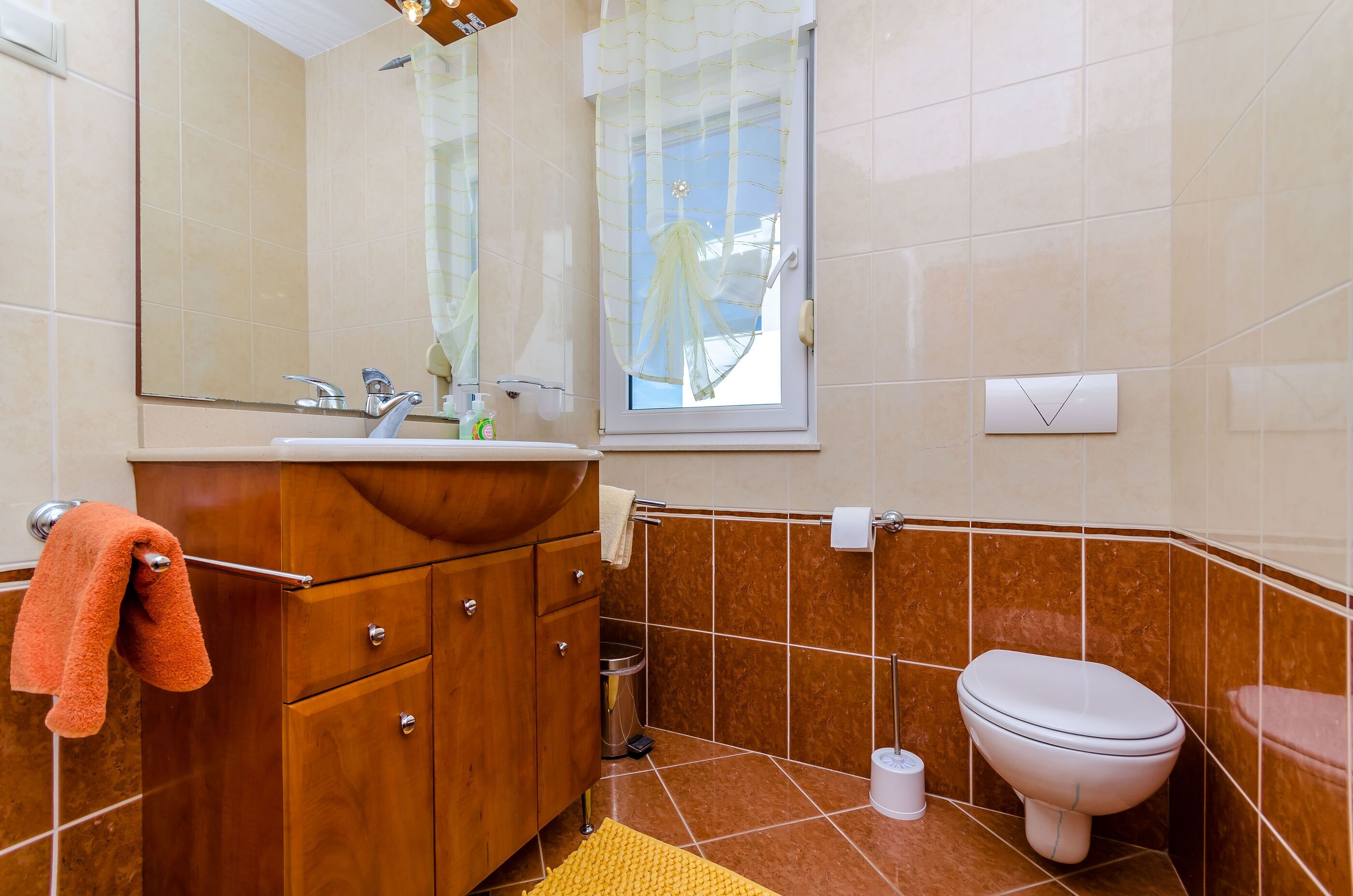A bathroom with a wooden vanity, a sink, a mirror, a toilet, and a window with a curtain.