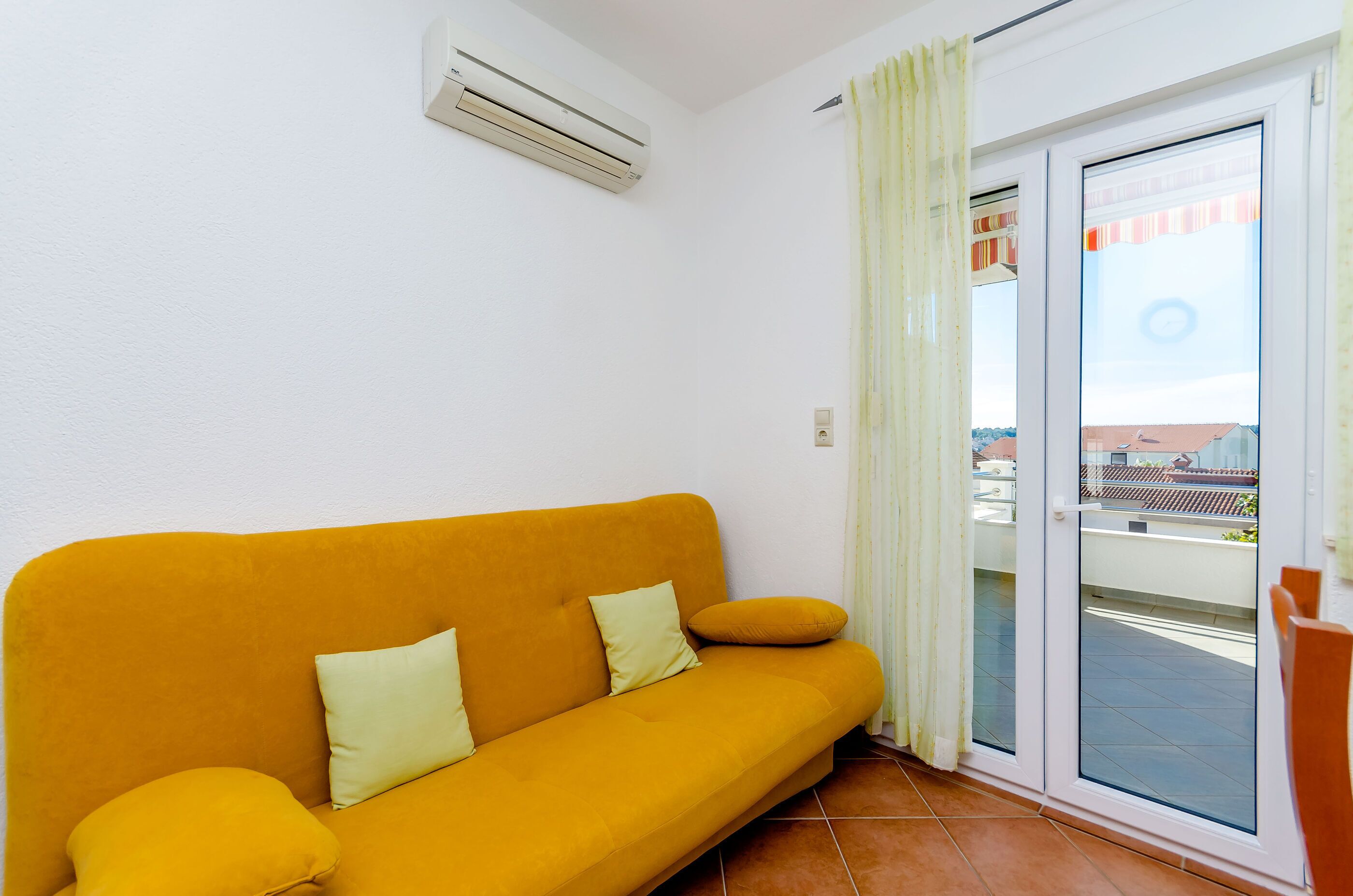 Een kamer met een gele bank, een schuifdeur naar een balkon en een airconditioning op de muur.