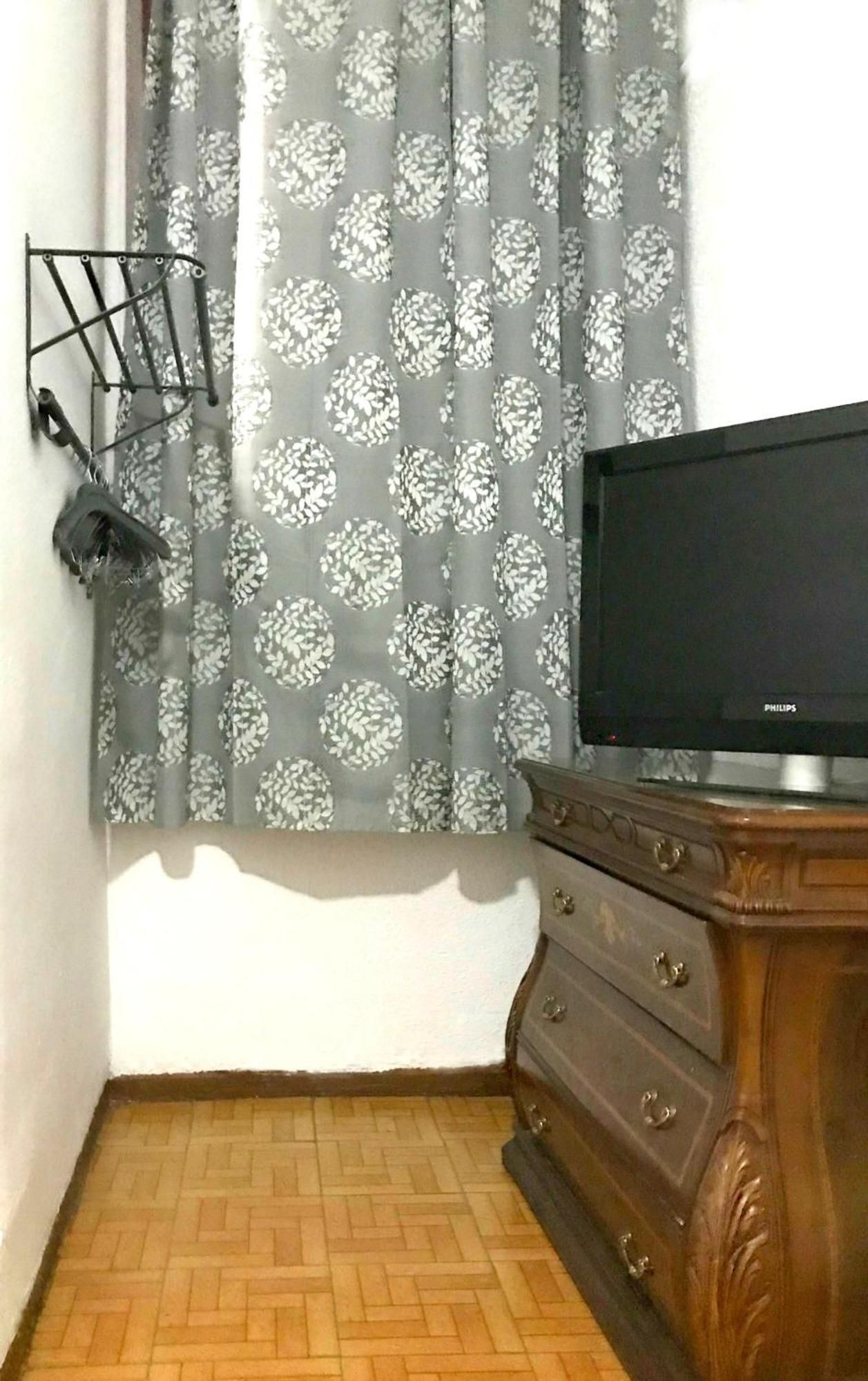Una habitación con una televisión sobre un aparador de madera, una ventana con cortinas estampadas y un zapatero en la pared.